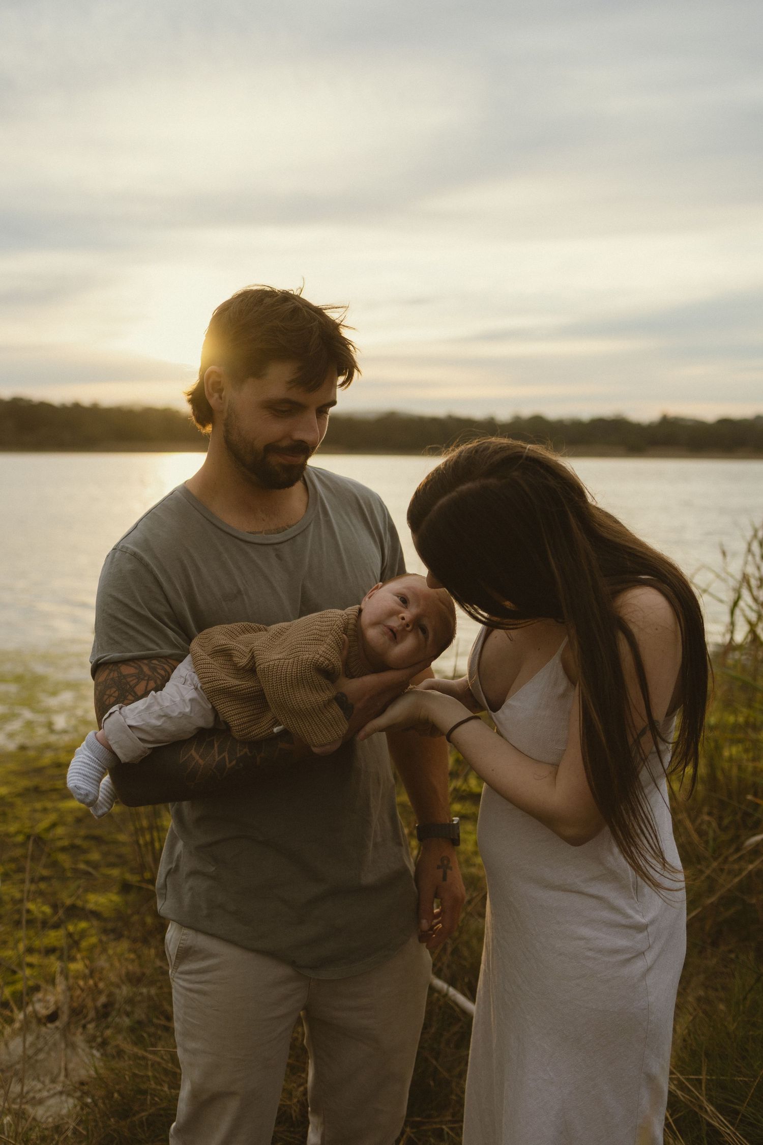 A couple admires their newborn baby outdoors by a lake at golden sunset, man with tattoos holding the infant.