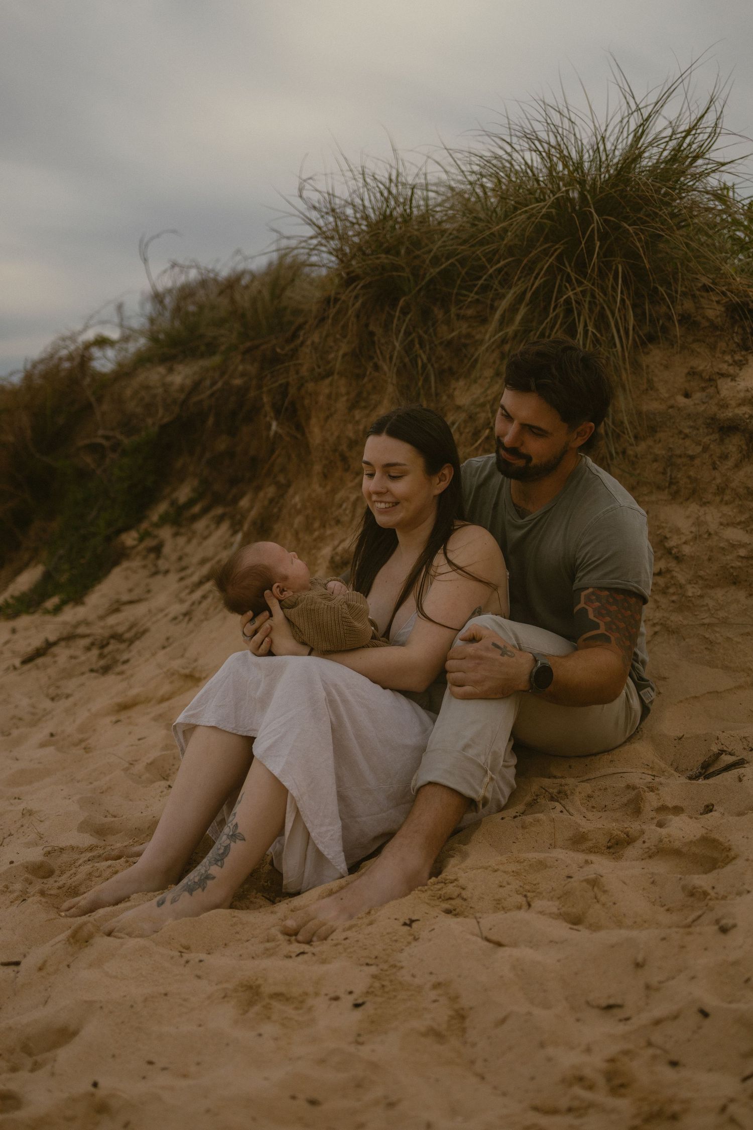 A couple sits on sandy beach dunes holding their newborn baby, surrounded by coastal grass under an overcast sky.