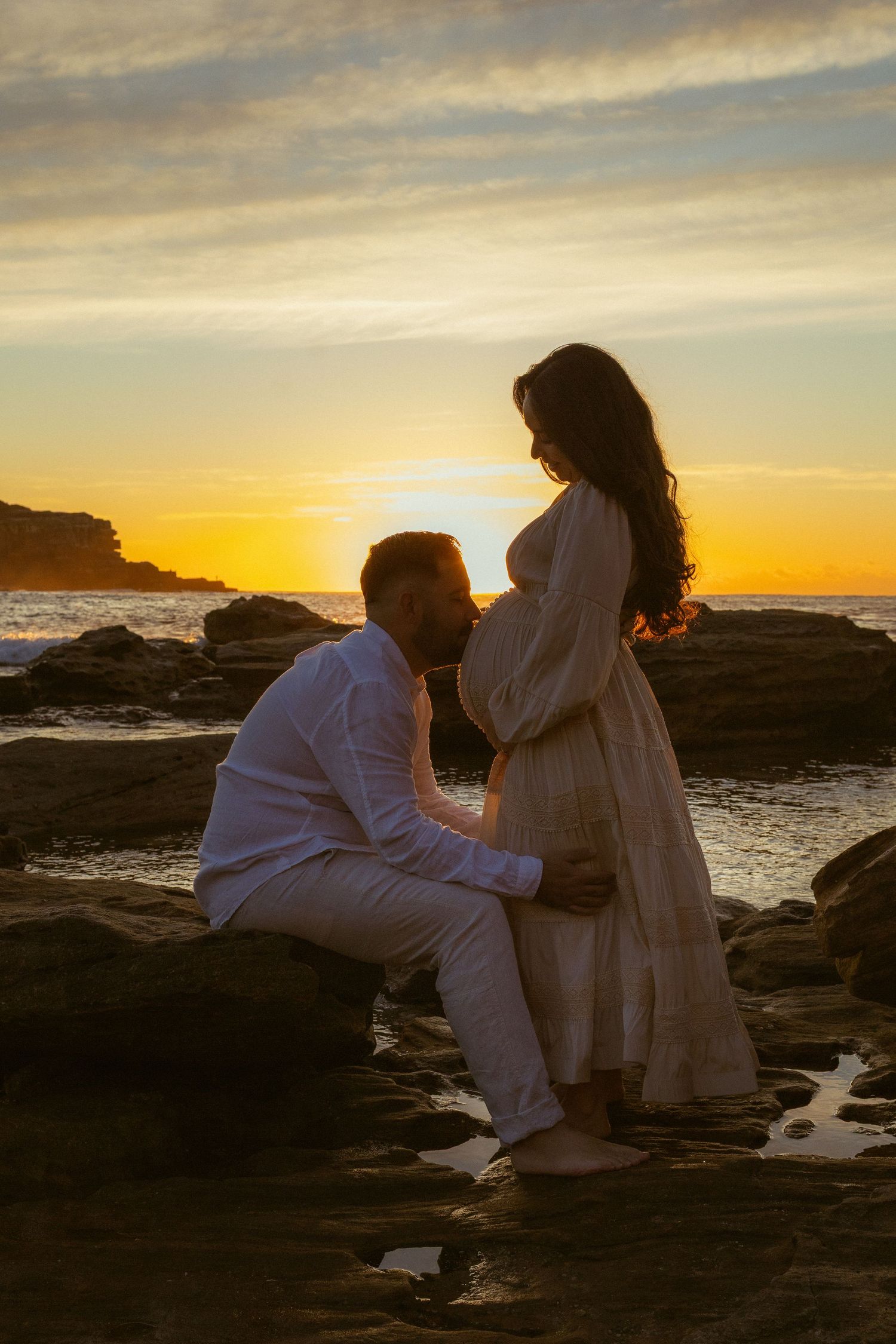 A man kneels before a pregnant woman on rocky beach at golden sunset, both dressed in white in a tender maternity photo.
