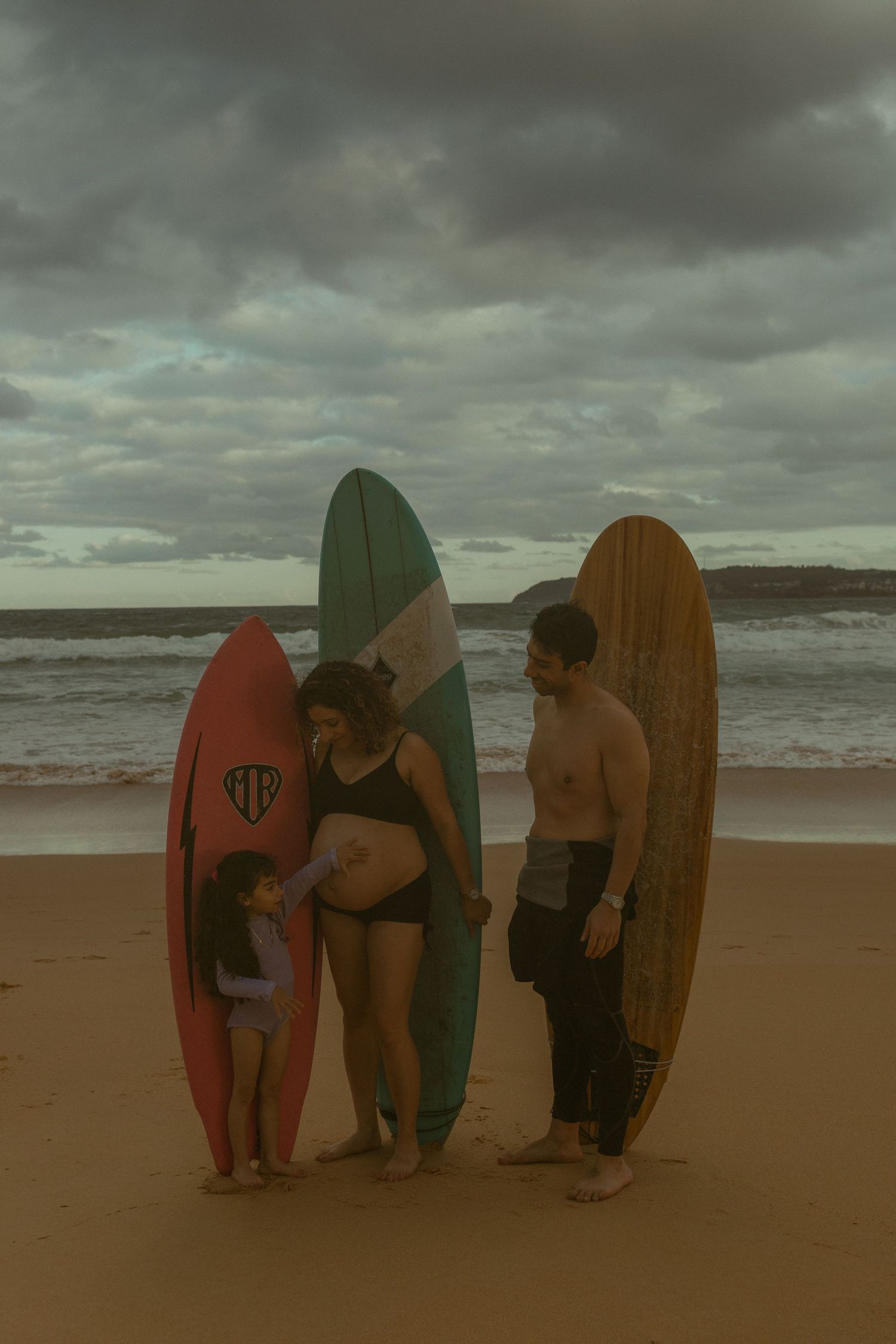 Pregnant woman, man, and child stand with colorful surfboards on a cloudy beach shore.