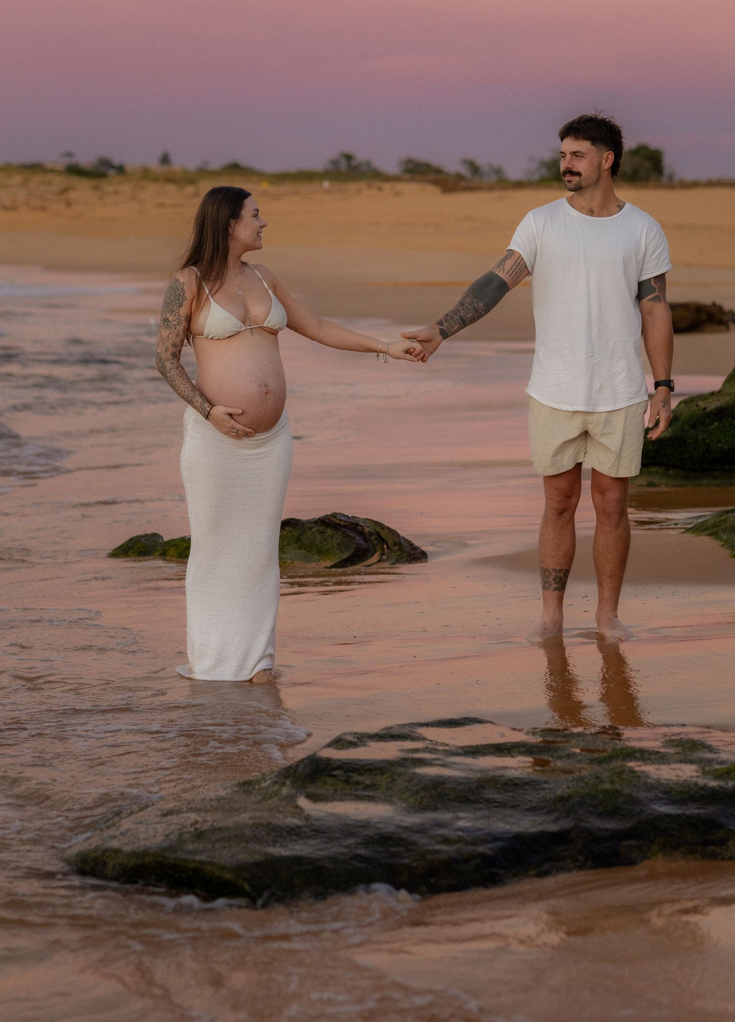 Pregnant couple holding hands on beach at sunset, woman in white dress showing baby bump, man in white shirt and shorts.