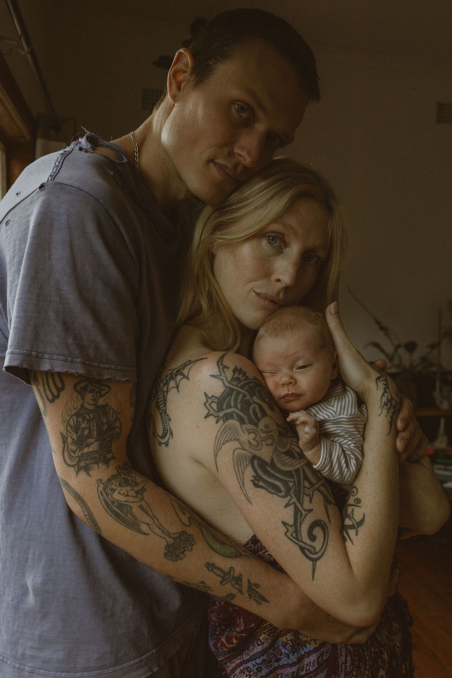 Tattooed couple tenderly holding their sleeping newborn baby in a warm, intimate home portrait.