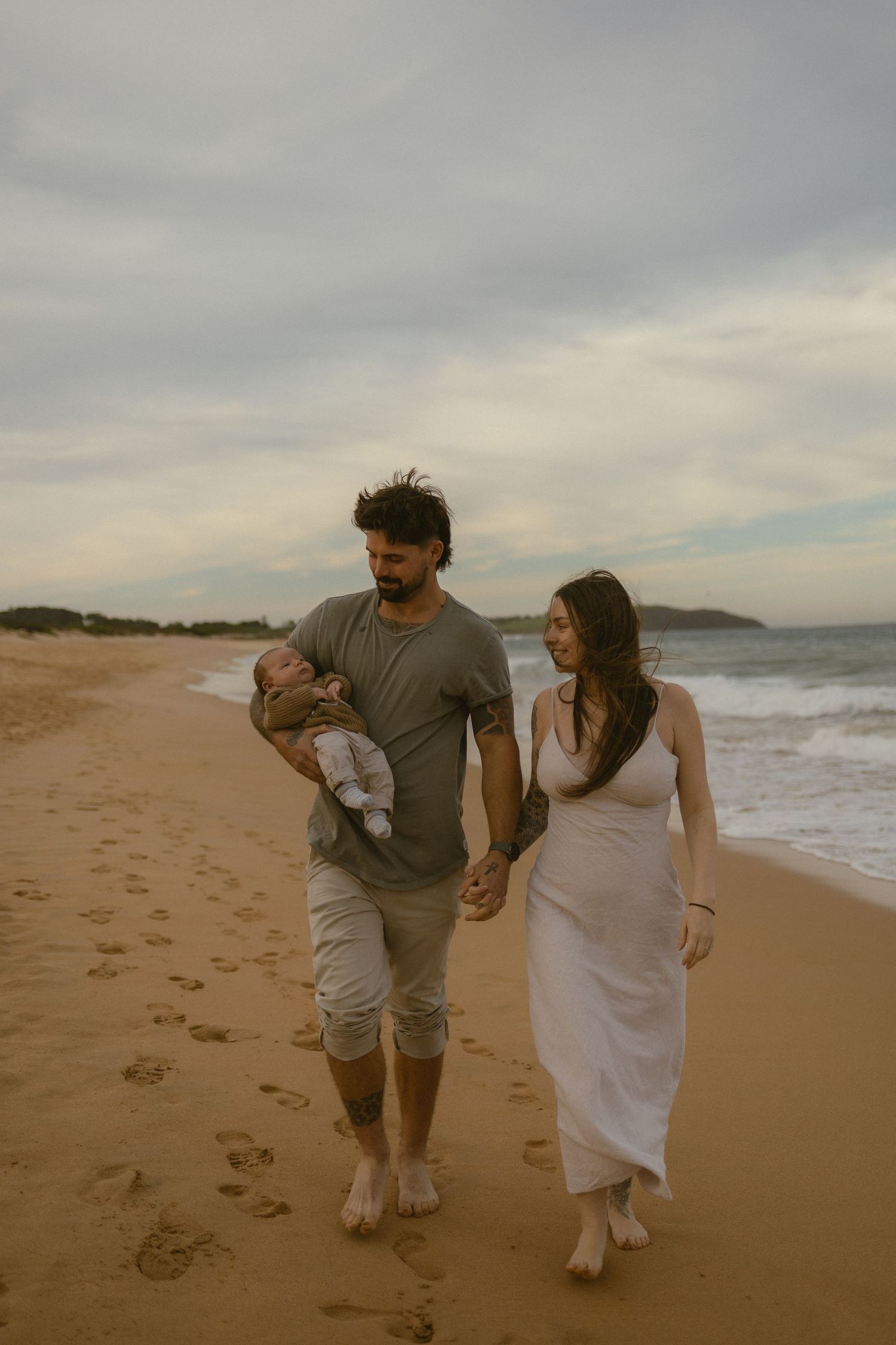 Young family walks barefoot on sandy beach at dusk, father holding newborn baby while holding mothers hand.