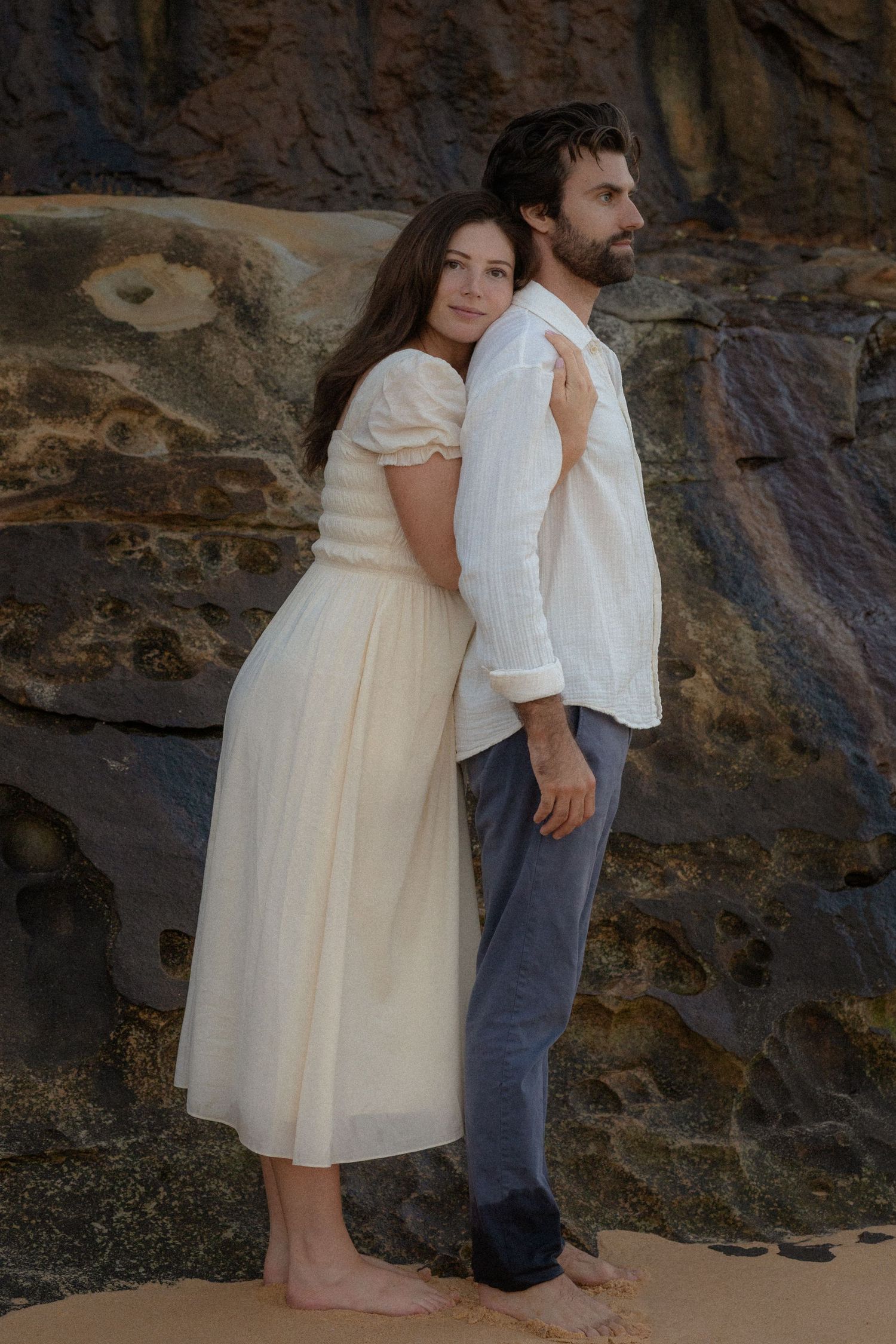 A couple in white attire poses barefoot against rocky backdrop, woman embracing man from behind in romantic outdoor setting.