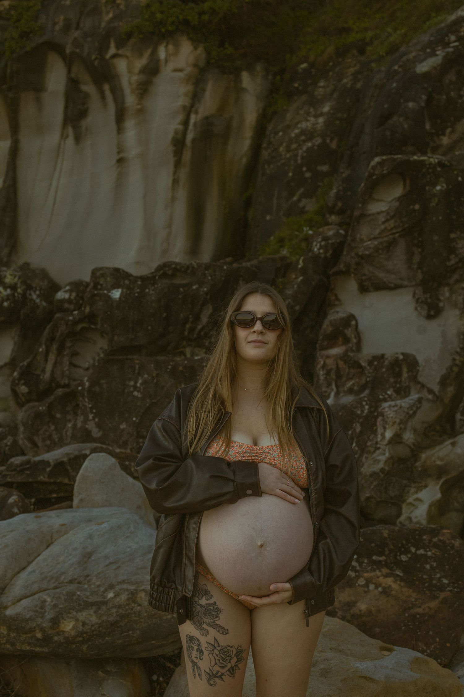Pregnant woman in sunglasses and dark jacket holds baby bump outdoors near rocky cliff backdrop.