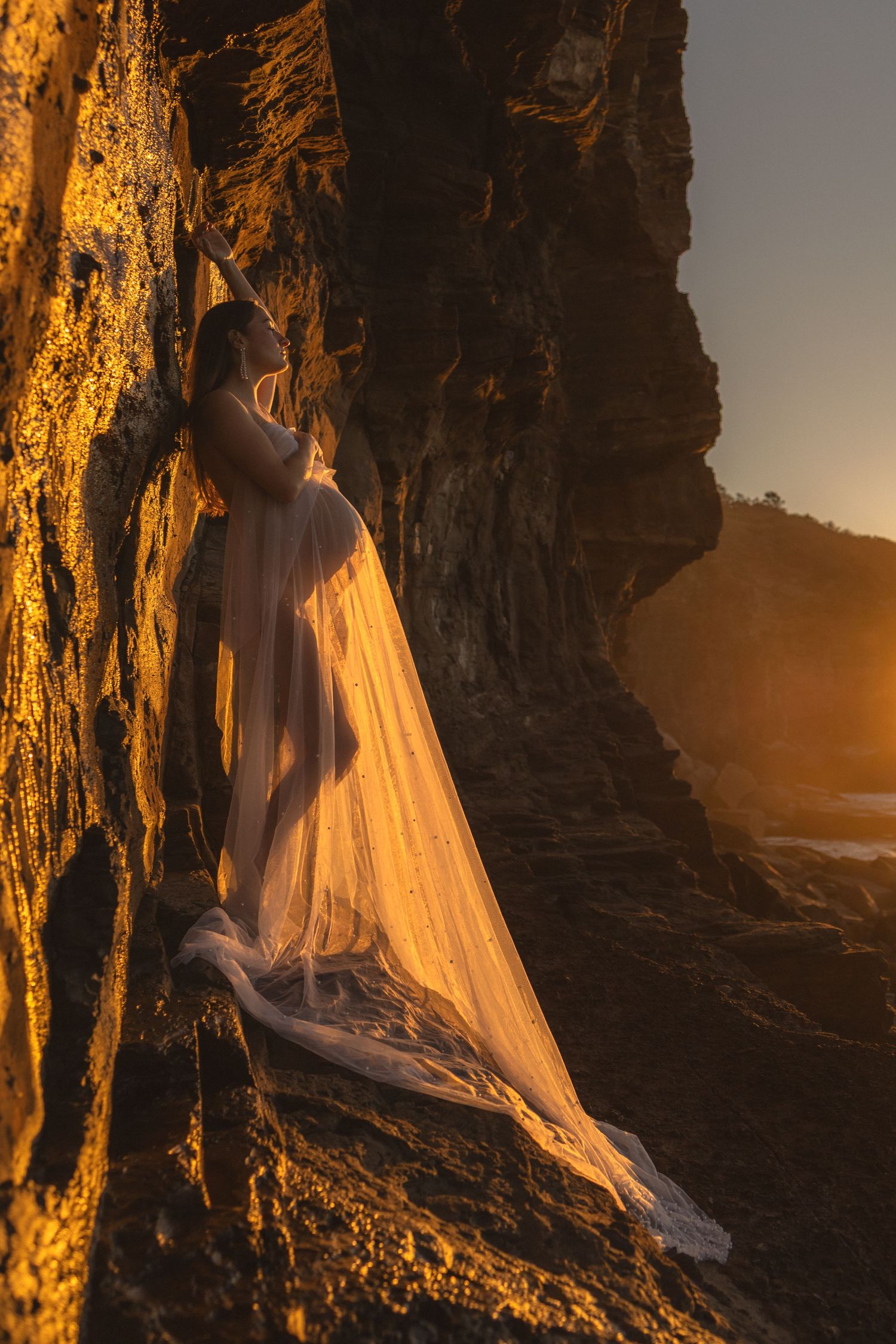 Pregnant woman in sheer white gown leaning against golden sunlit coastal rocks at sunset.