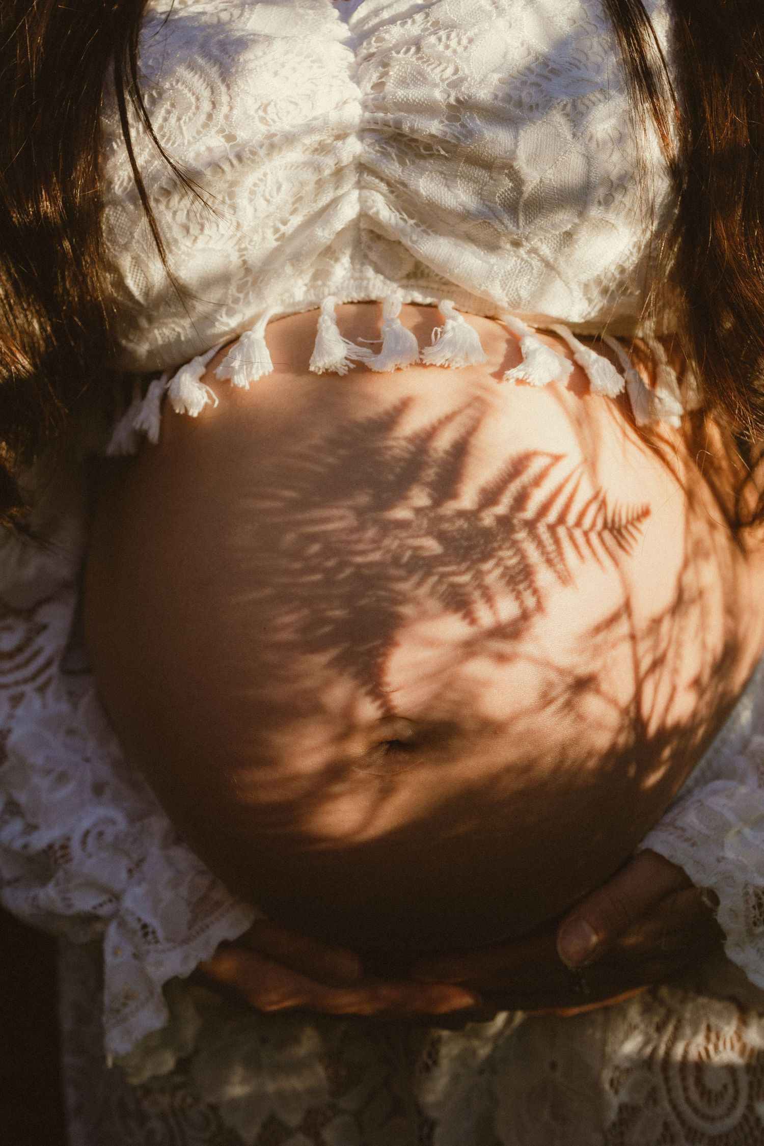 Close-up of a pregnant woman's bare belly with fern leaf shadows in warm sunlight, wearing a white lace dress.