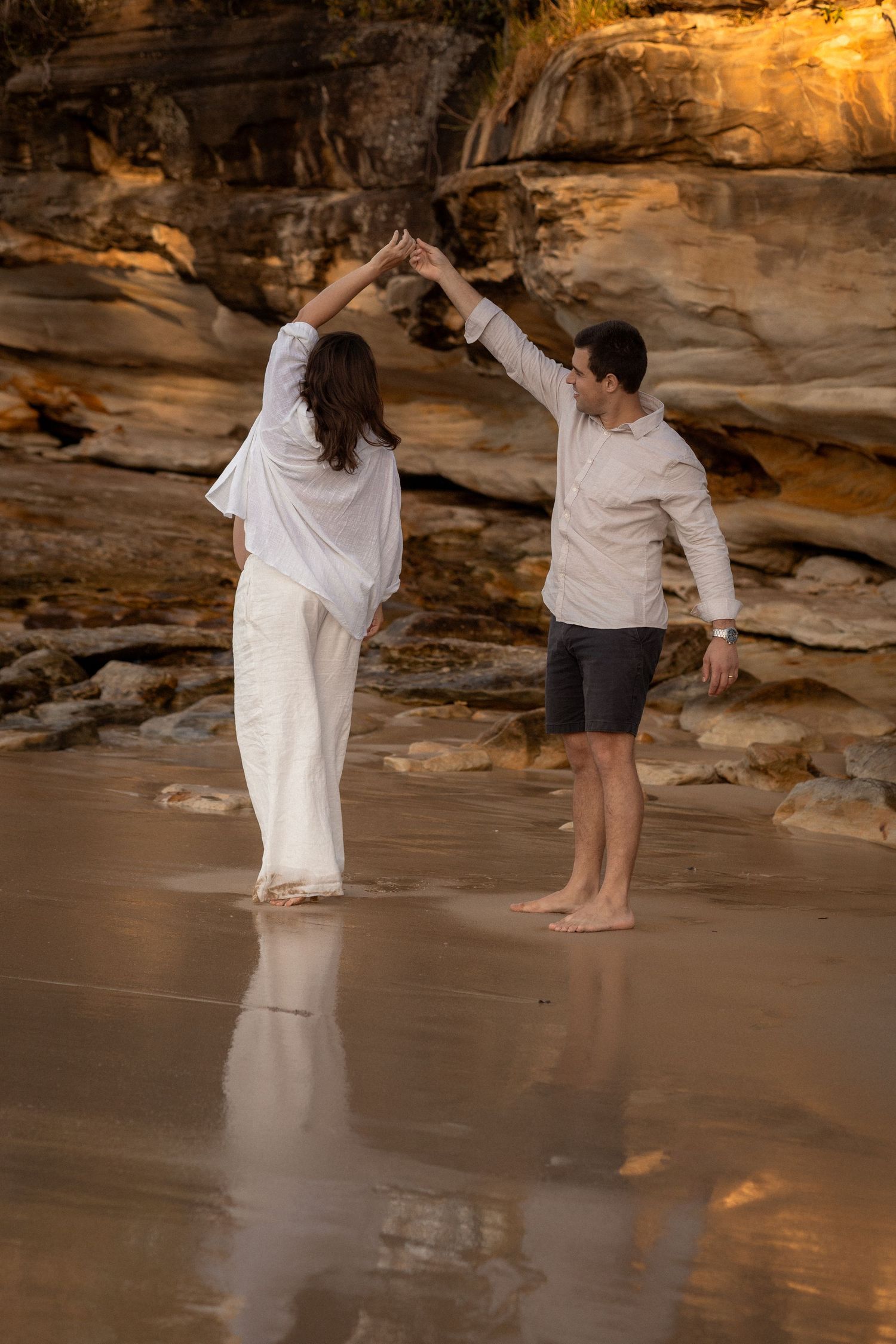 Two people in white and dark clothing walk along a sandy beach near rocky cliffs at sunset.