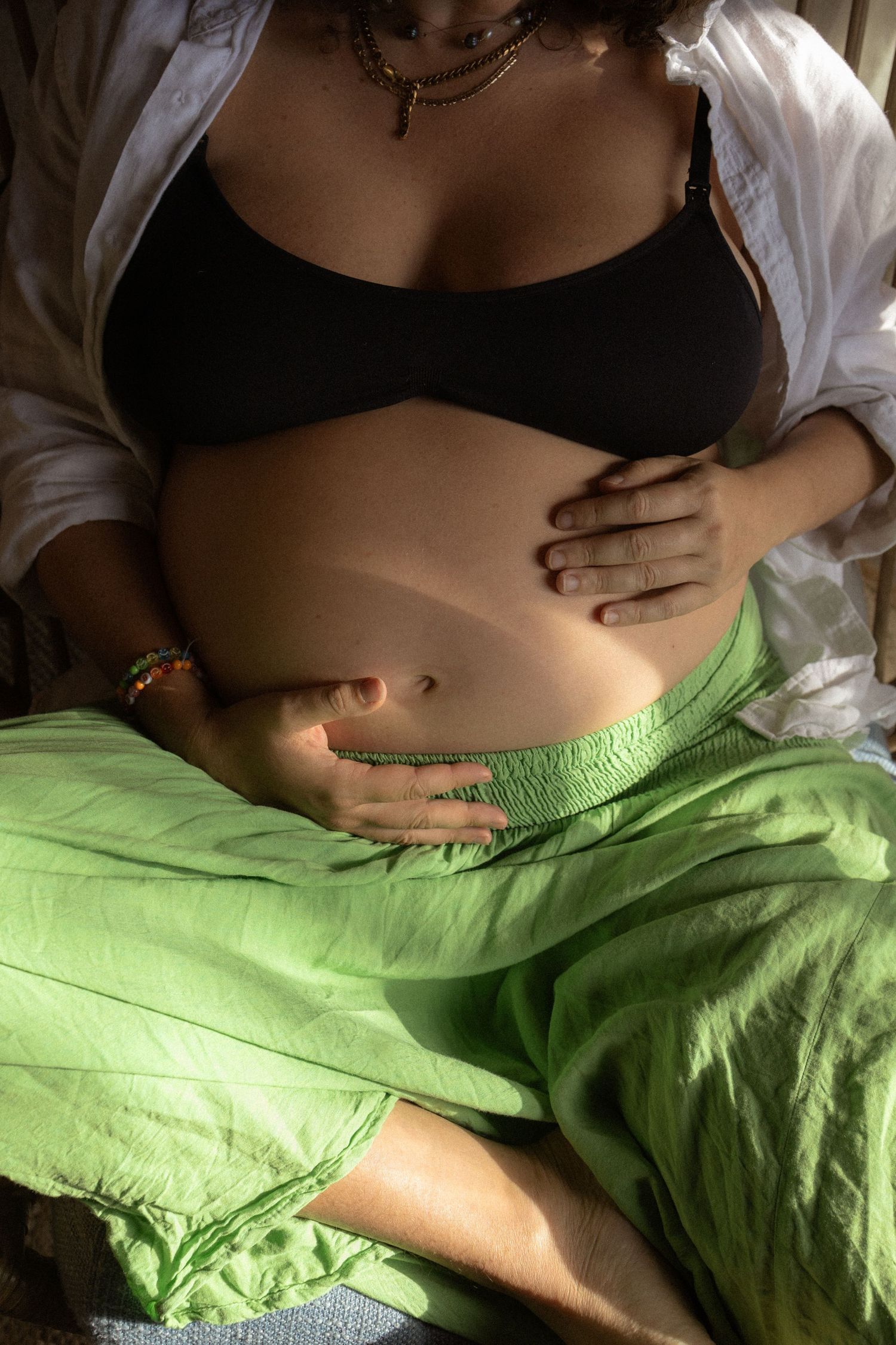 Pregnant woman in black bralette and green pants cradling her bare baby bump in warm sunlight.