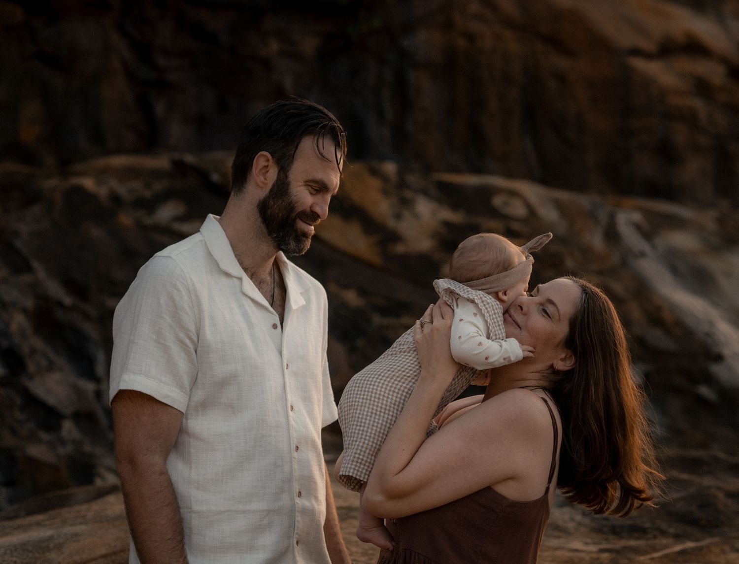 A couple shares a tender moment with their baby outdoors at sunset against rocky cliffs.