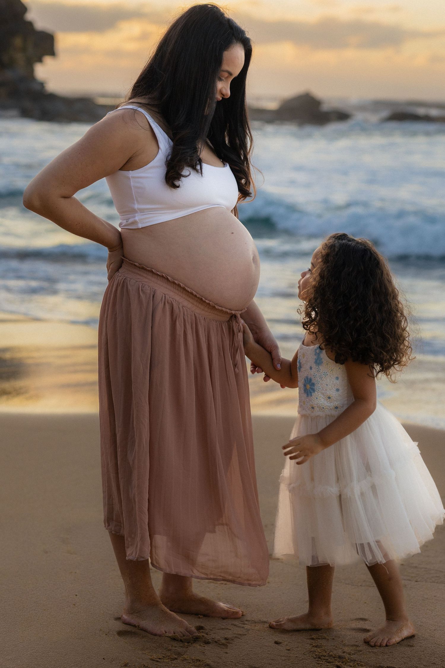 A tender moment captured at sunset on a beach with a parent and child in matching flowy dresses against ocean waves.