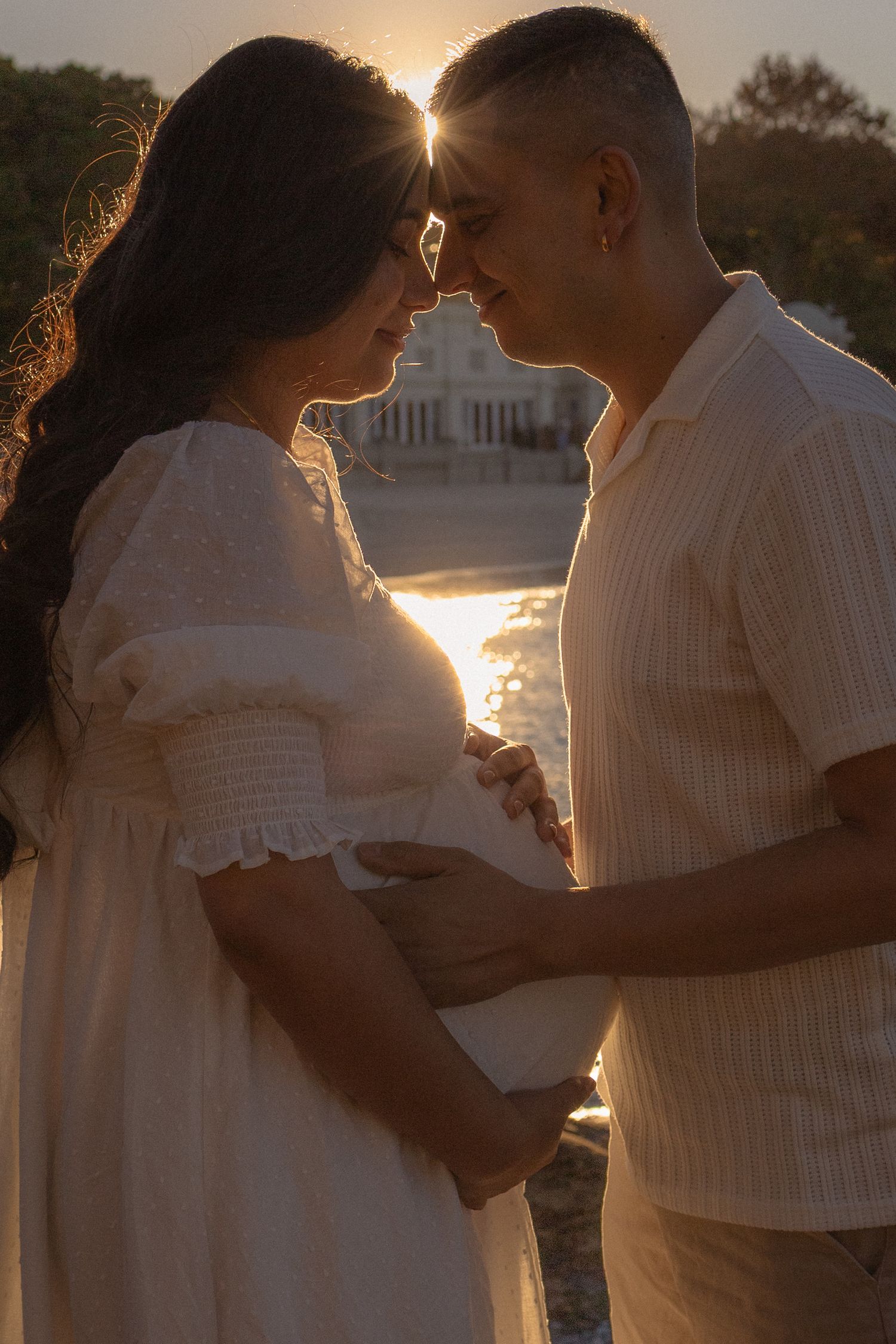 Couple sharing a tender moment at sunset, man holding pregnant woman's belly in golden backlit glow.