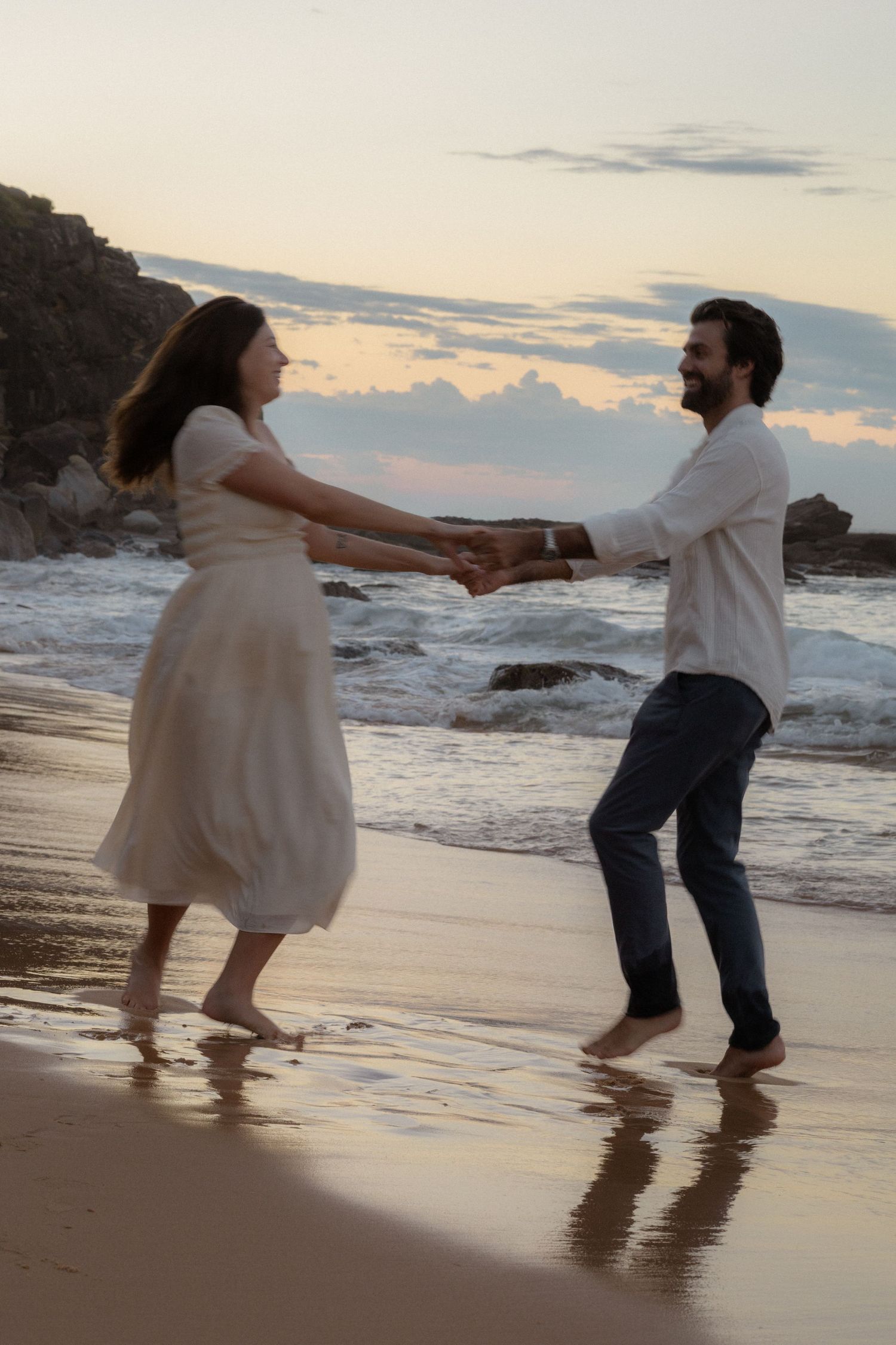 Couple dancing barefoot on a sandy beach at sunset, holding hands near ocean waves.