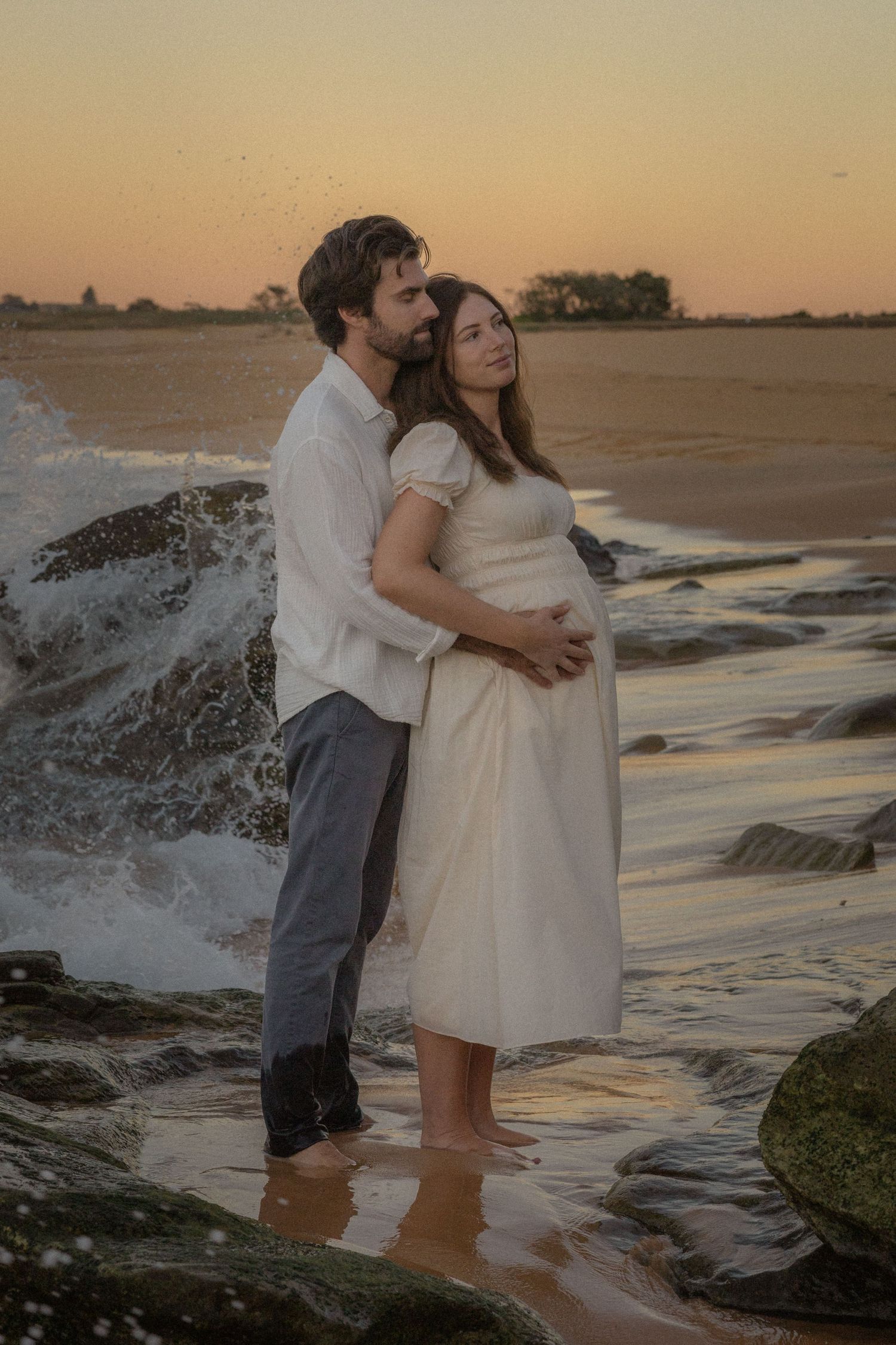 Couple embracing at sunset on rocky beach, pregnant woman in white dress, waves crashing behind them.