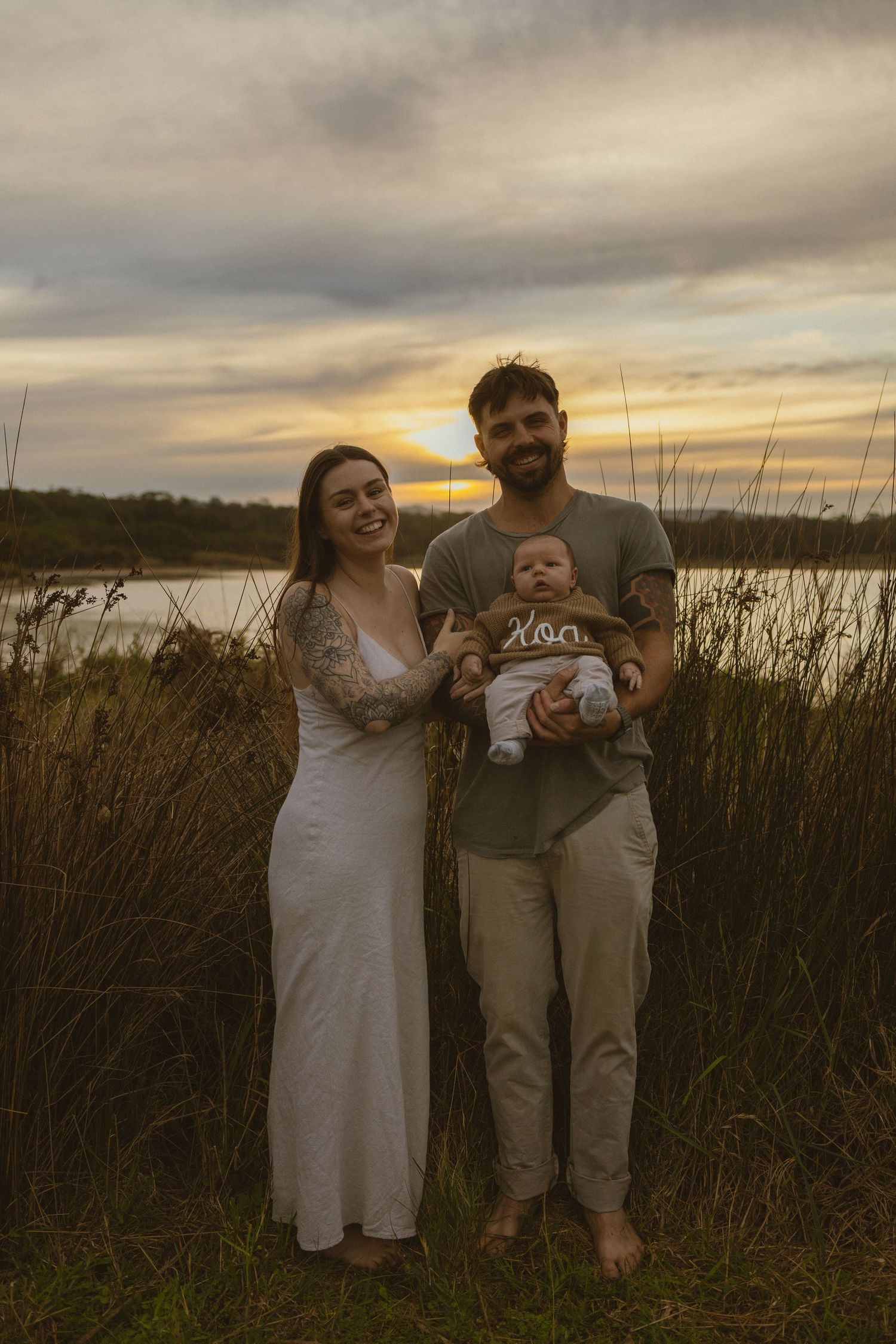 Young family with baby poses at golden sunset near wetlands, woman in white dress and tattooed man holding infant.