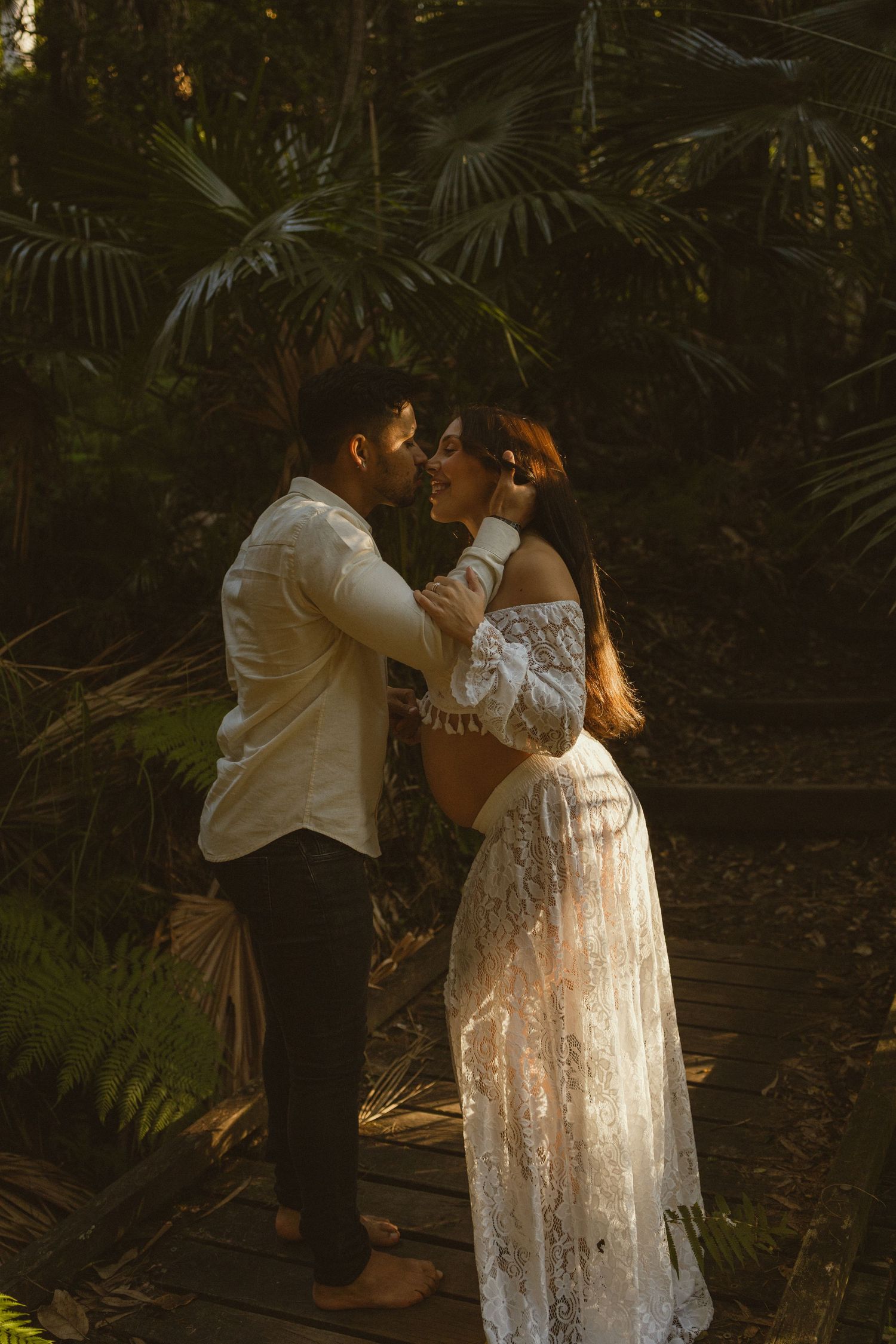 A couple shares a romantic moment in a lush tropical forest, the woman wearing an elegant white lace dress.