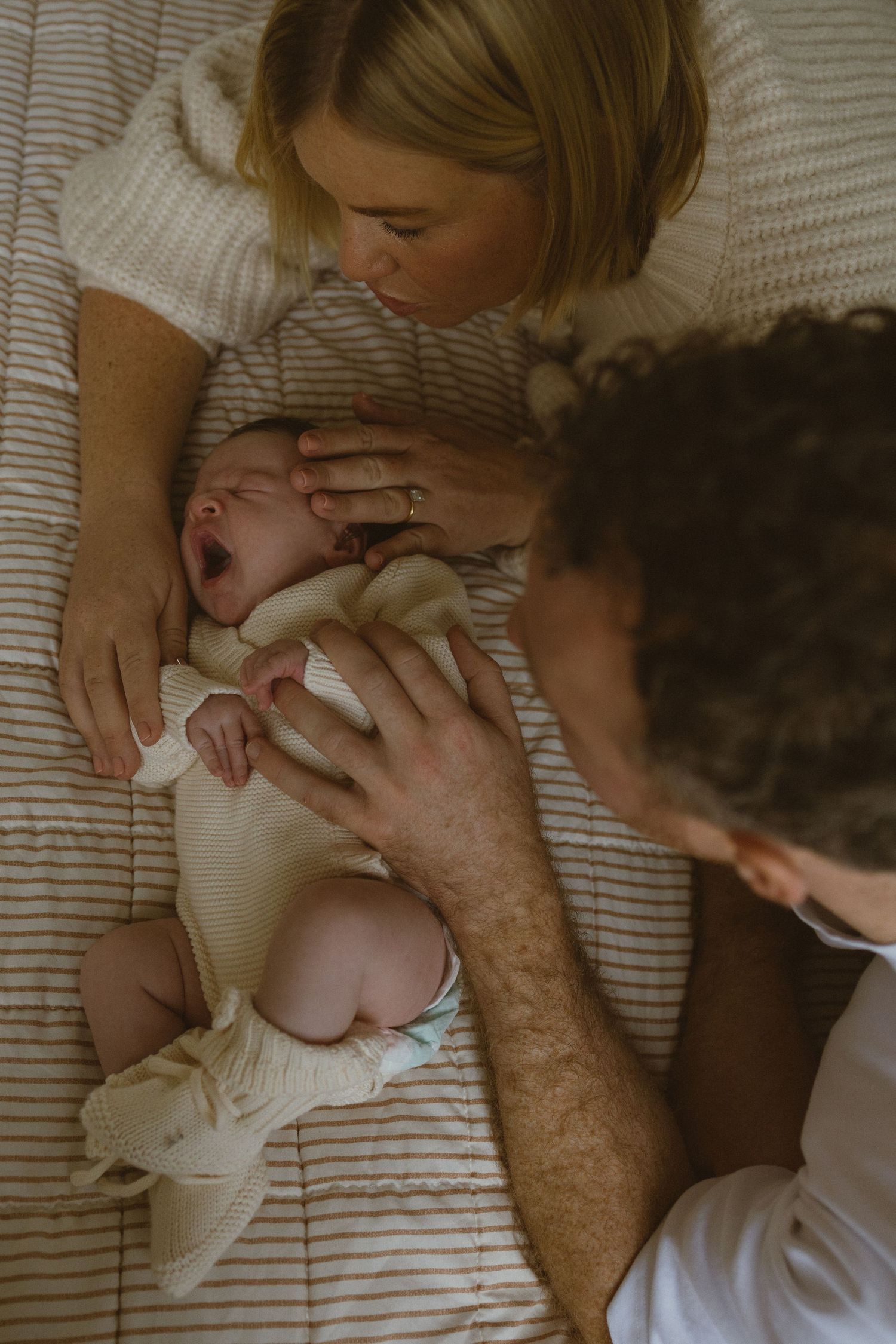 Parents lovingly gaze at their yawning newborn baby lying on a striped bed.