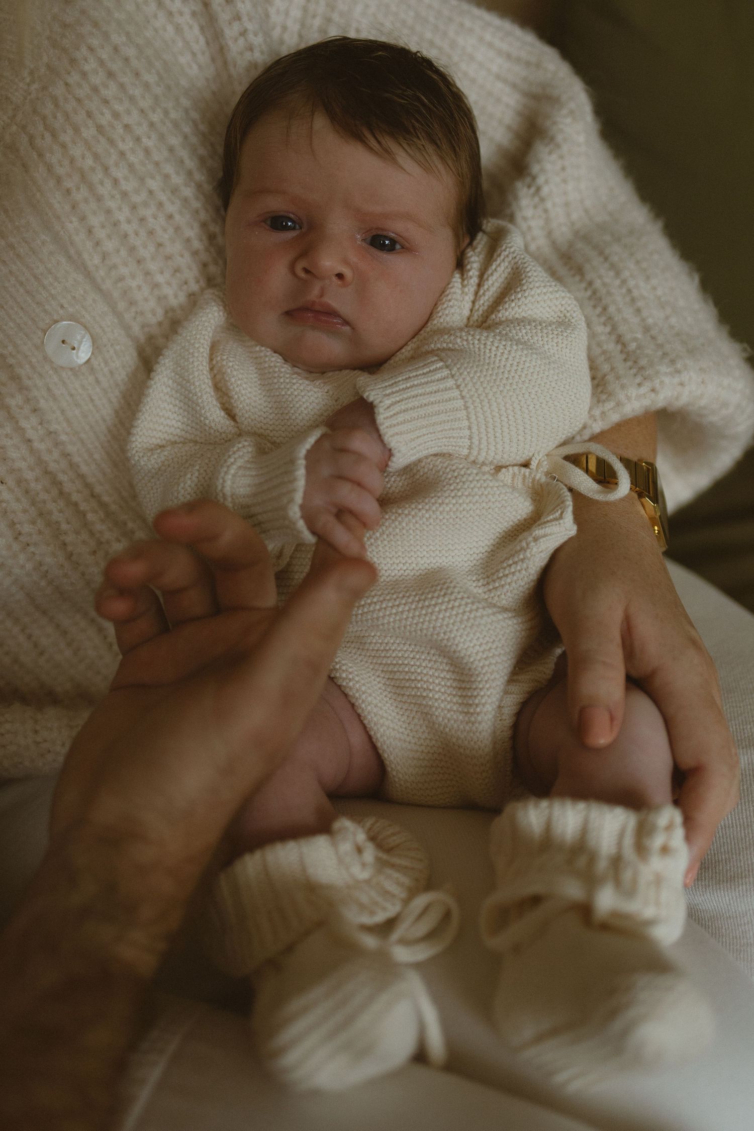 Newborn baby in cream knit romper and booties being cradled in adult hands, looking alert and peaceful.