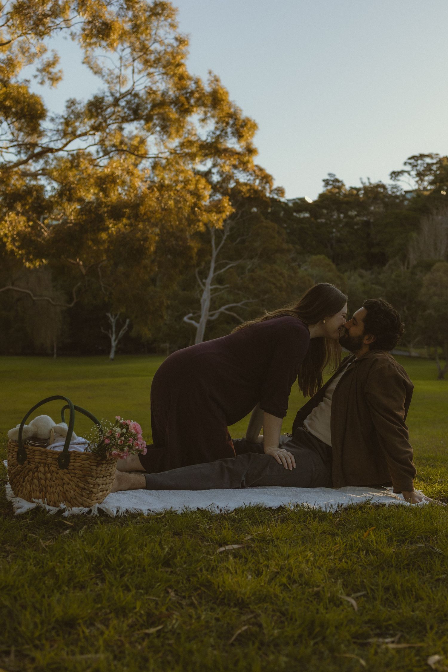 Couple sharing a romantic kiss during a sunset picnic on a blanket in a lush green park with autumn trees.