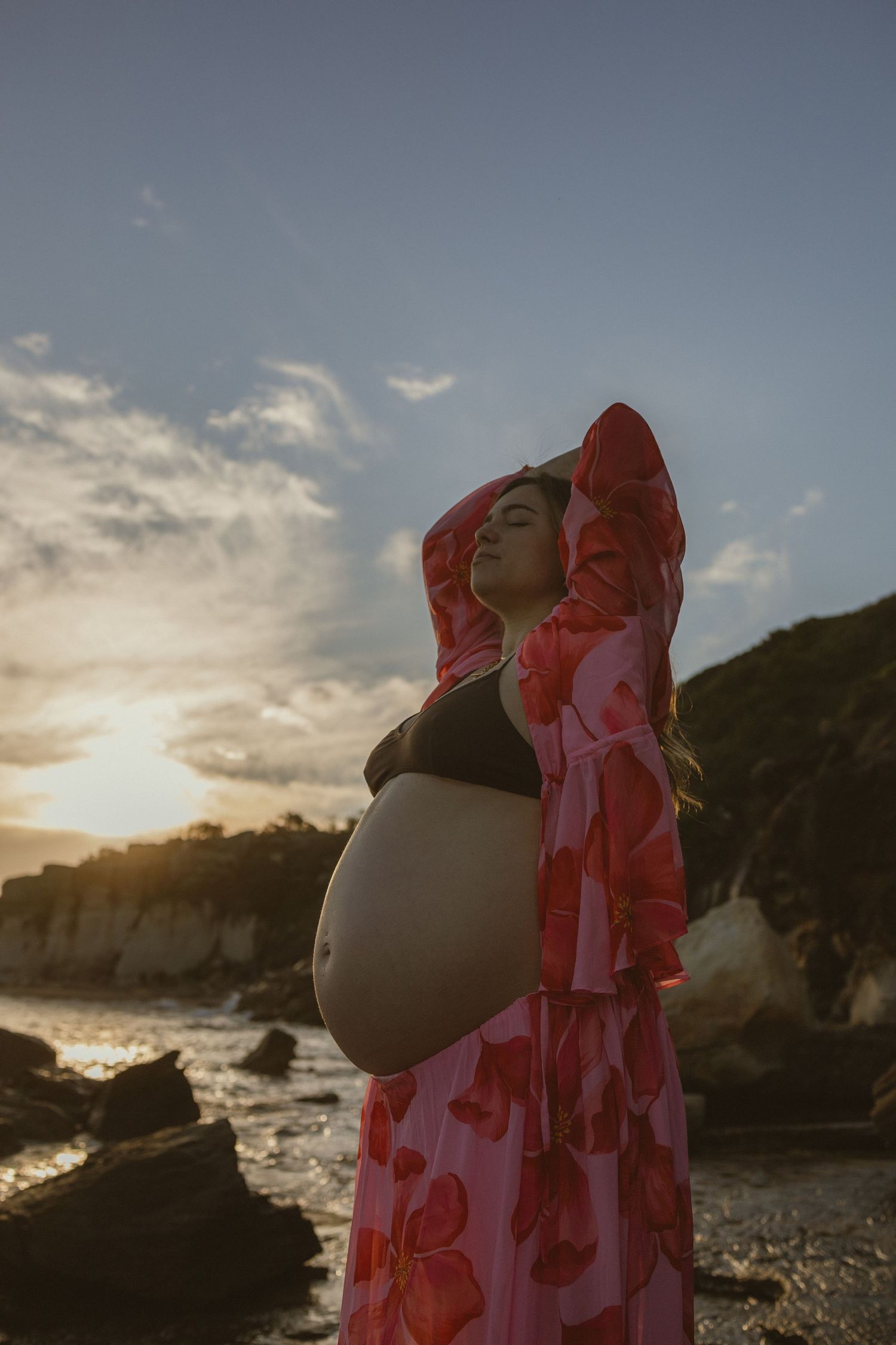 Pregnant woman in pink floral robe poses on rocky beach at sunset, hands raised, eyes closed, belly exposed.