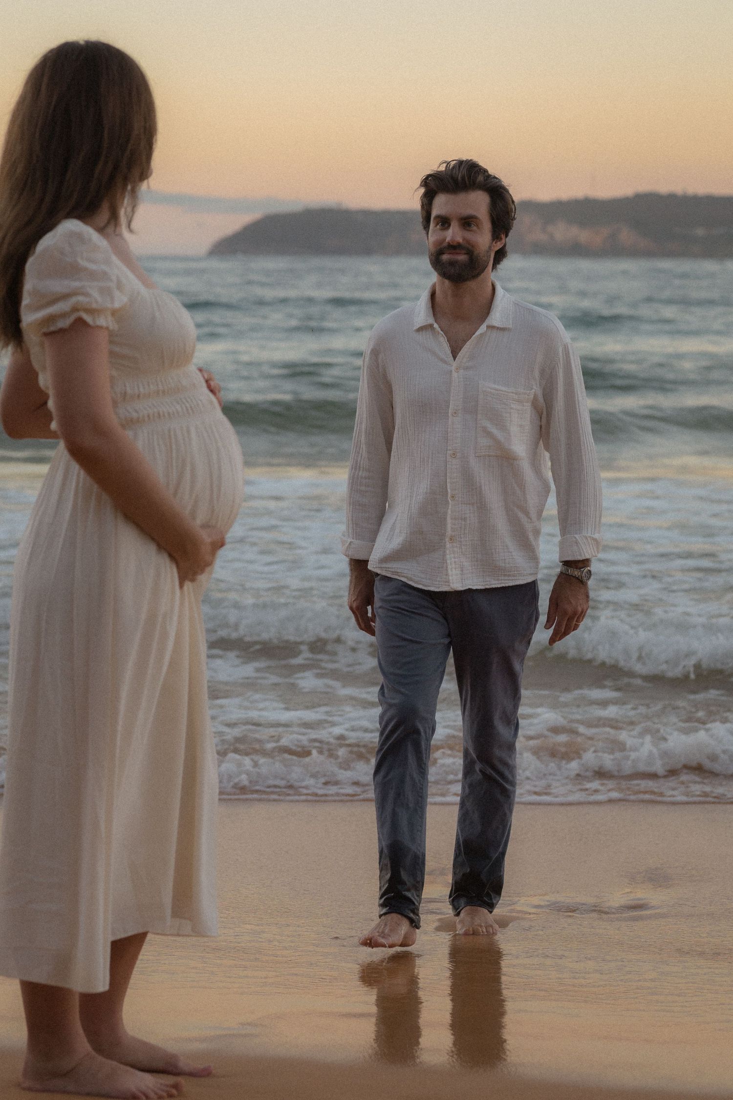 Pregnant woman in white dress stands with a man in white shirt on a beach at sunset.