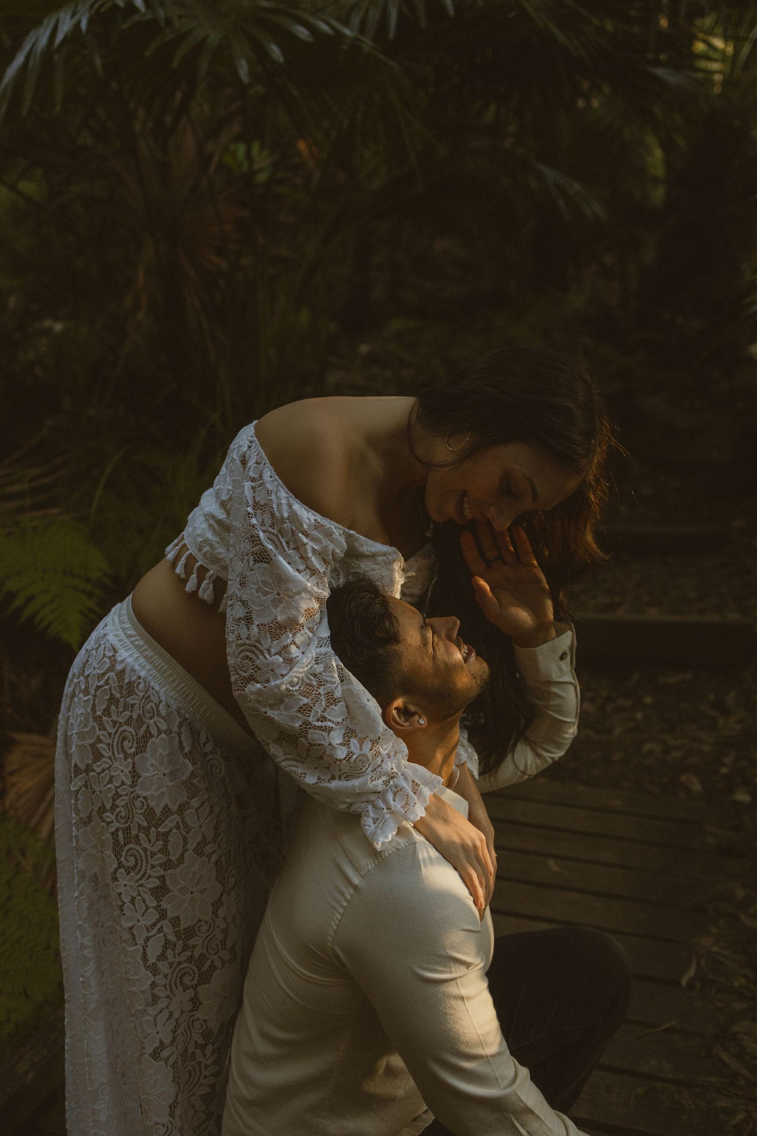 Bride in white lace gown leaning over groom in sunlit tropical garden during romantic wedding shoot.
