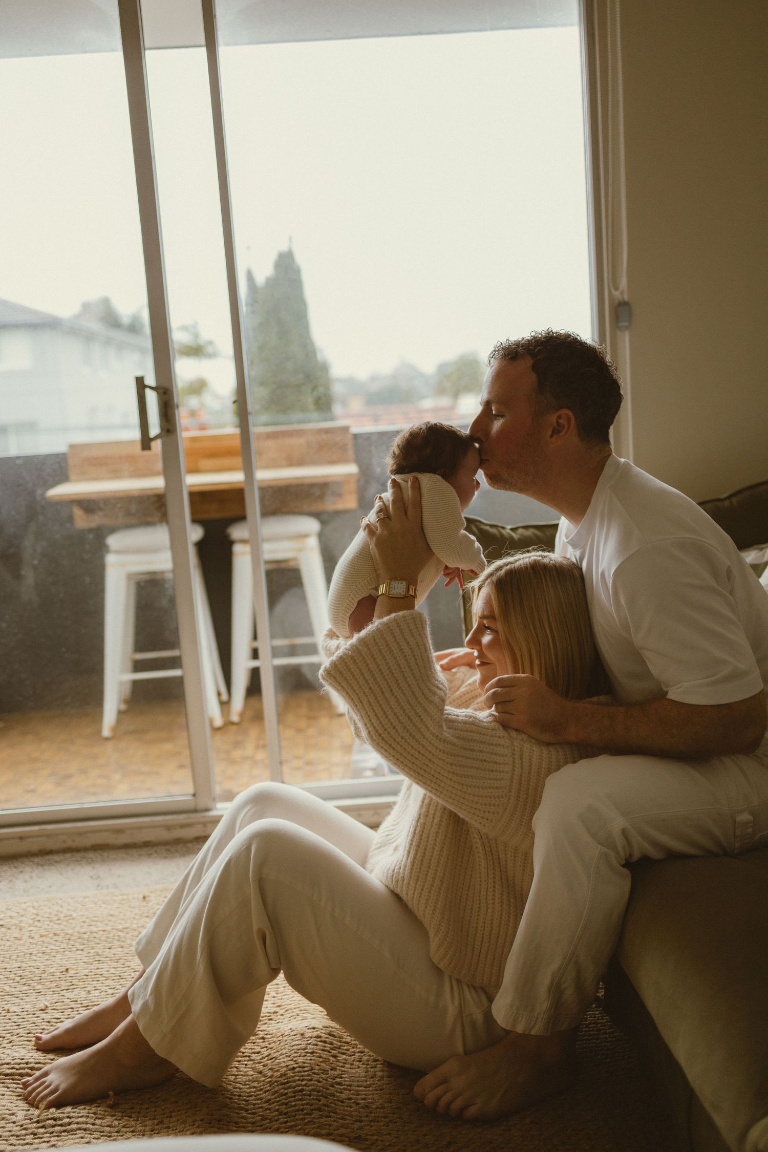 A couple sits on the floor by a sunny window, with the man kissing their newborn baby while the woman leans against him.