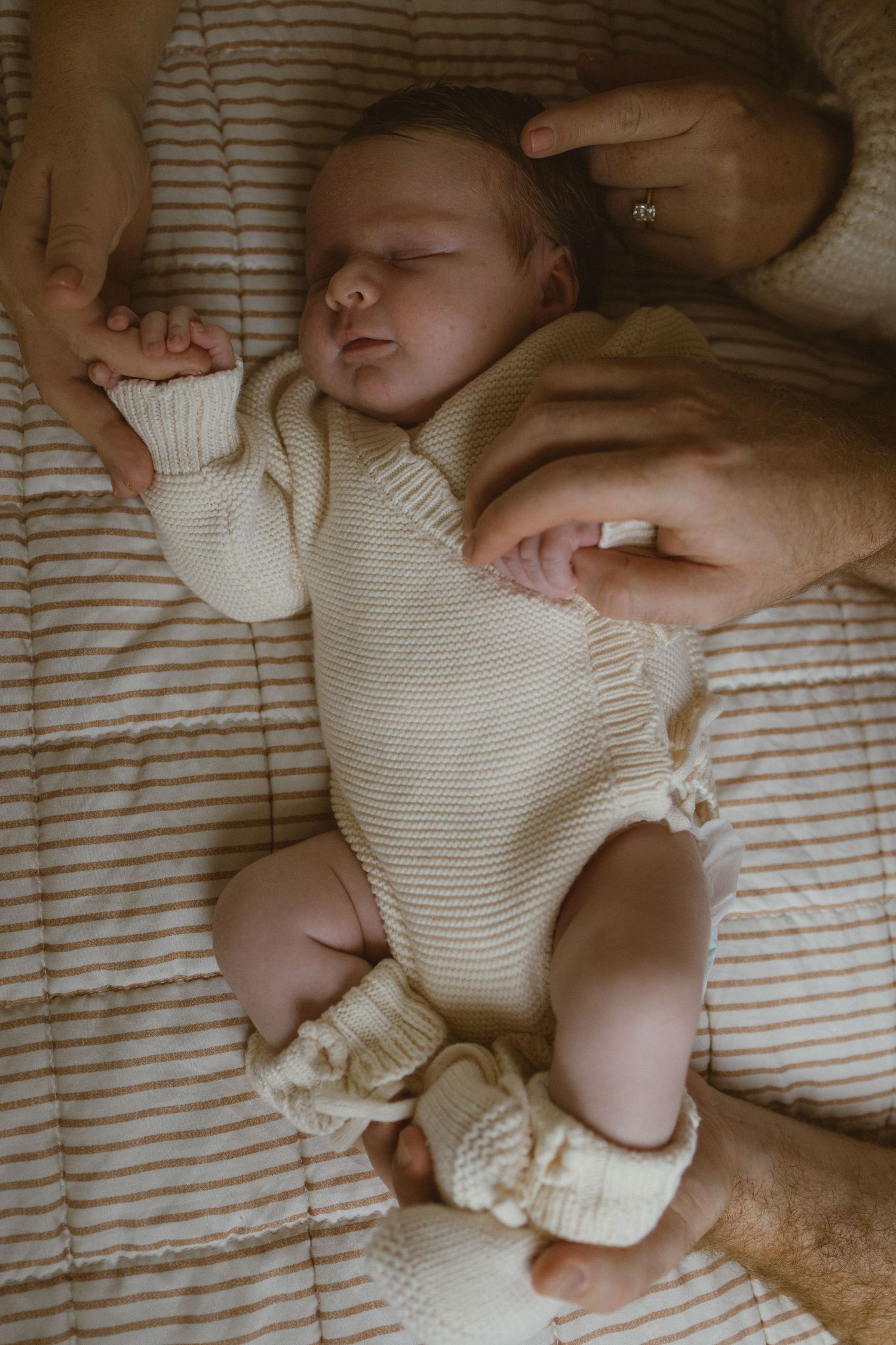 Newborn baby in cream knit romper and booties held gently by parents hands on striped bedding.