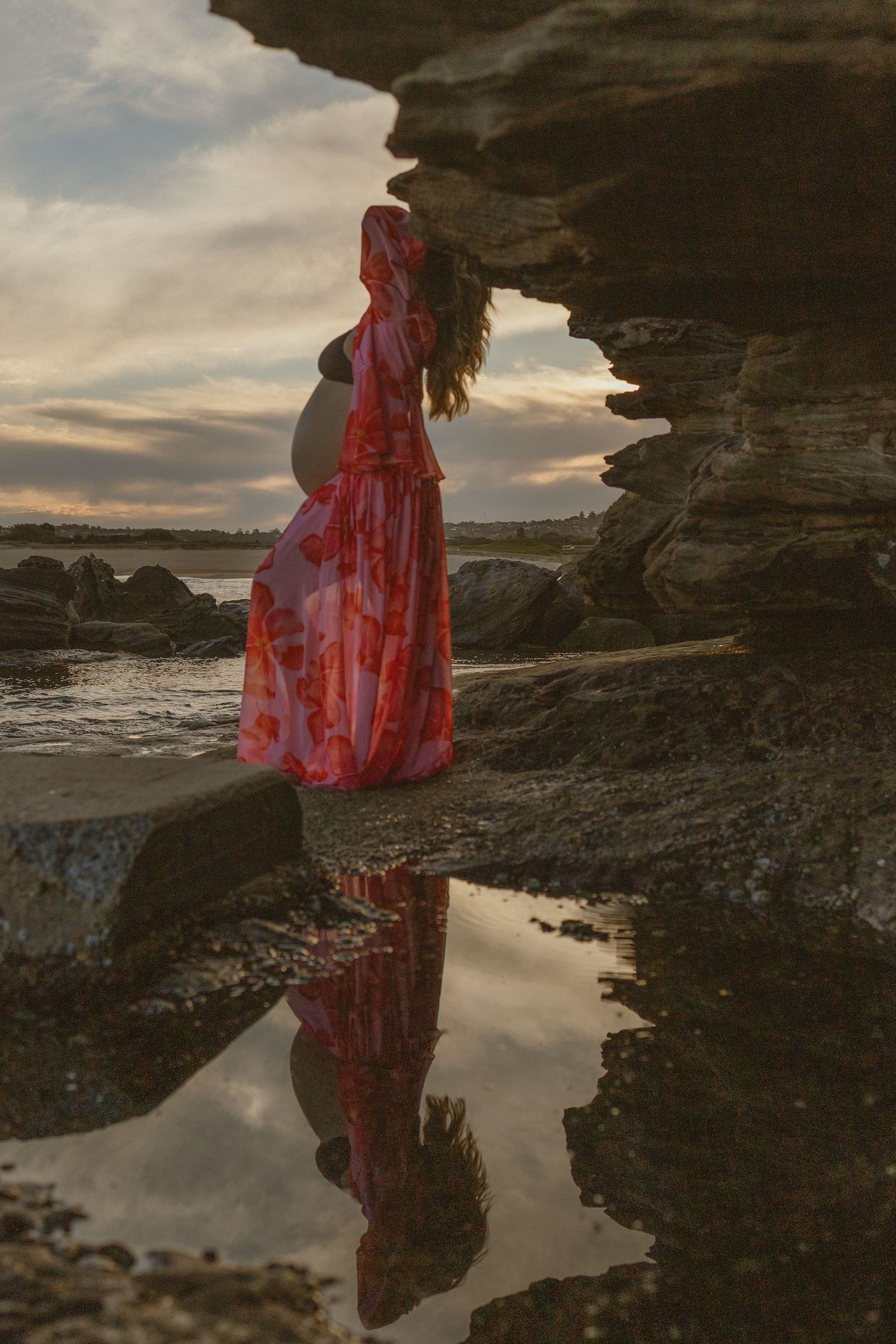 Pregnant woman in red floral dress stands on rocks reflected in tide pool at sunset.
