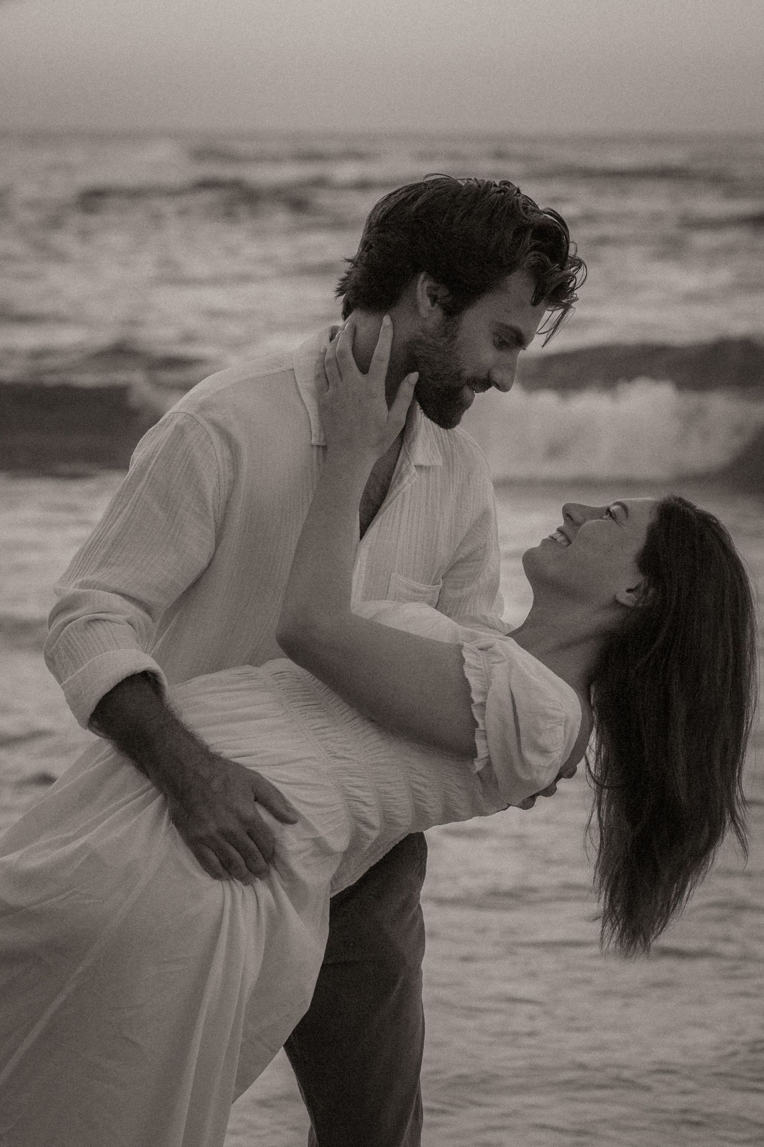 Black and white photo of a romantic couple embracing on a beach at sunset, waves crashing behind them.