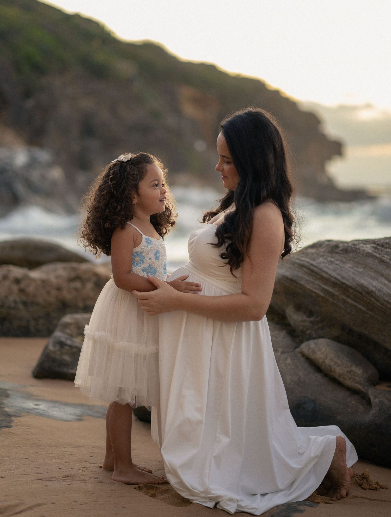 Two people in white dresses share tender moments on a rocky beach at sunset.