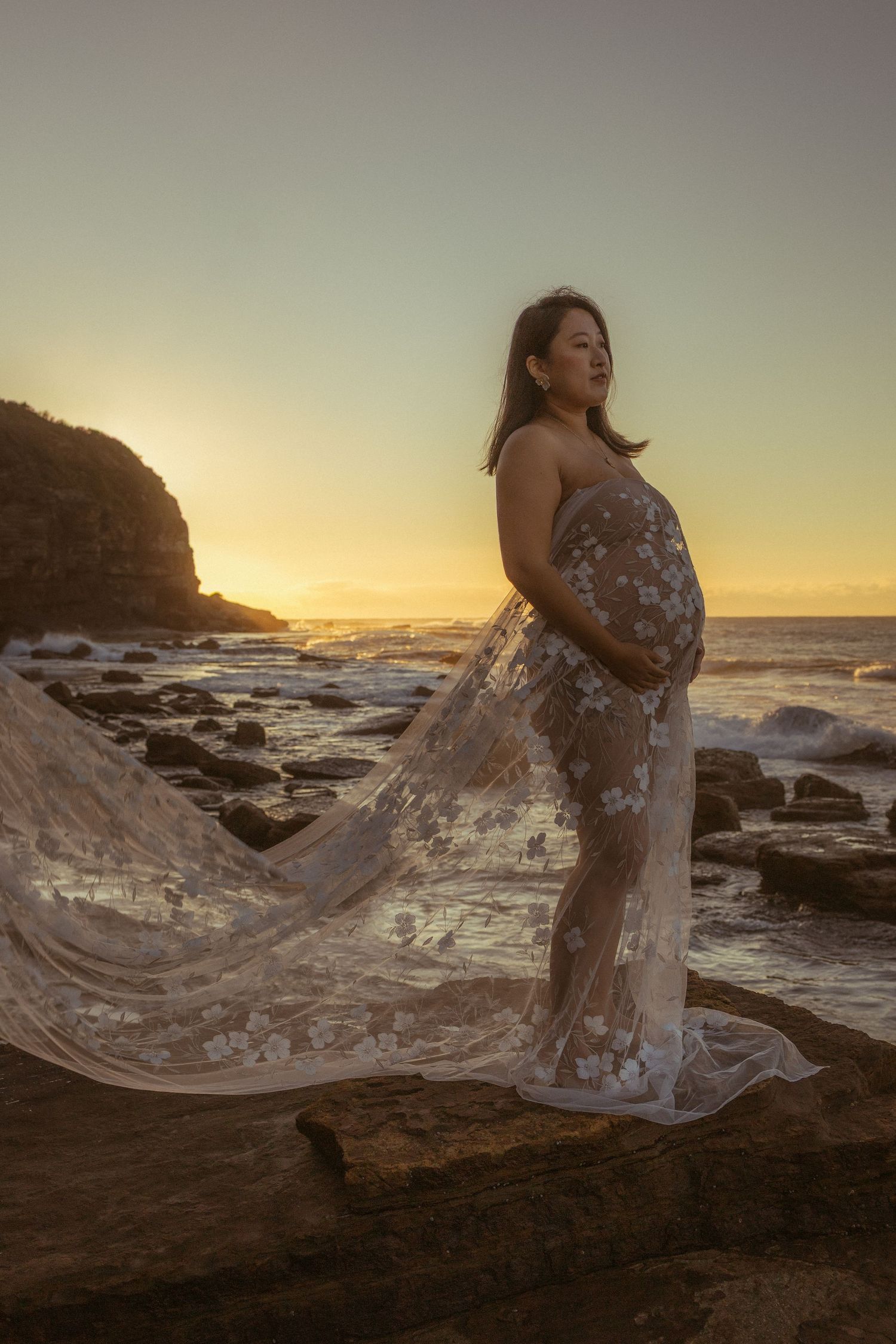 Pregnant woman in flowing lace gown stands on coastal rocks at golden sunset, cradling her baby bump.