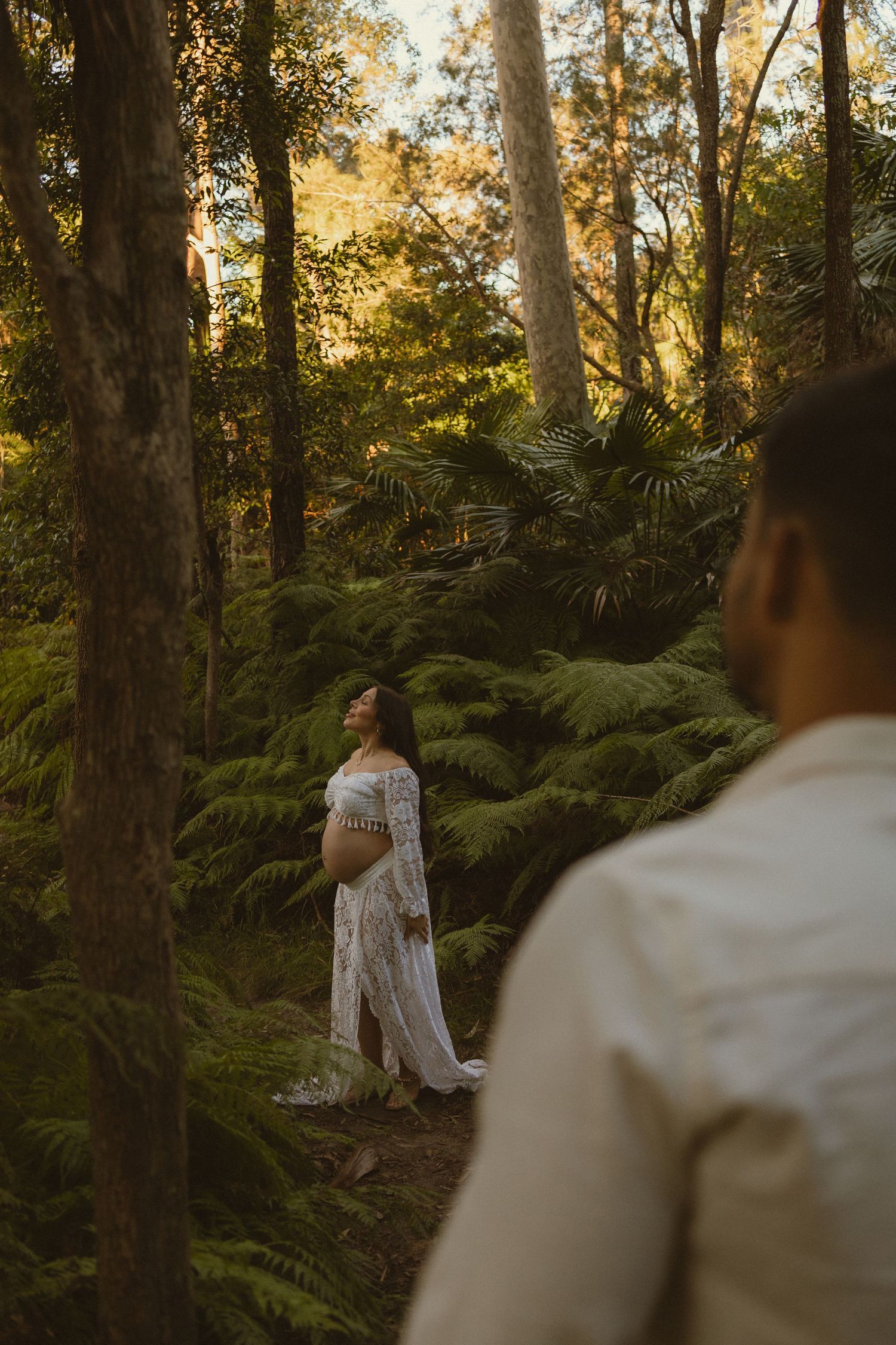 Pregnant woman in white lace dress standing peacefully among ferns in golden forest light, partner watching nearby.