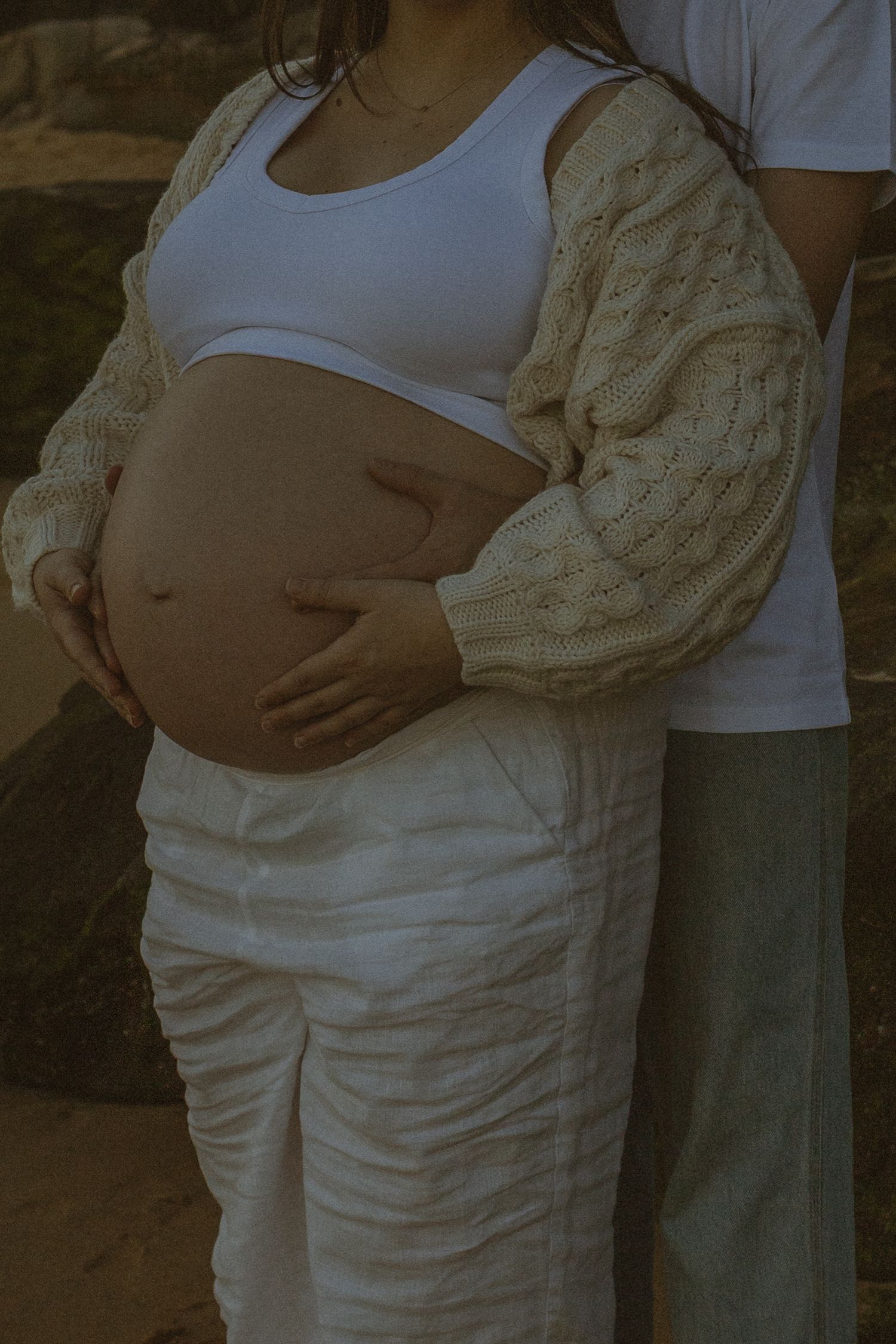Pregnant woman in white crop top and pants with partner's hand on baby bump, maternity photo shoot.