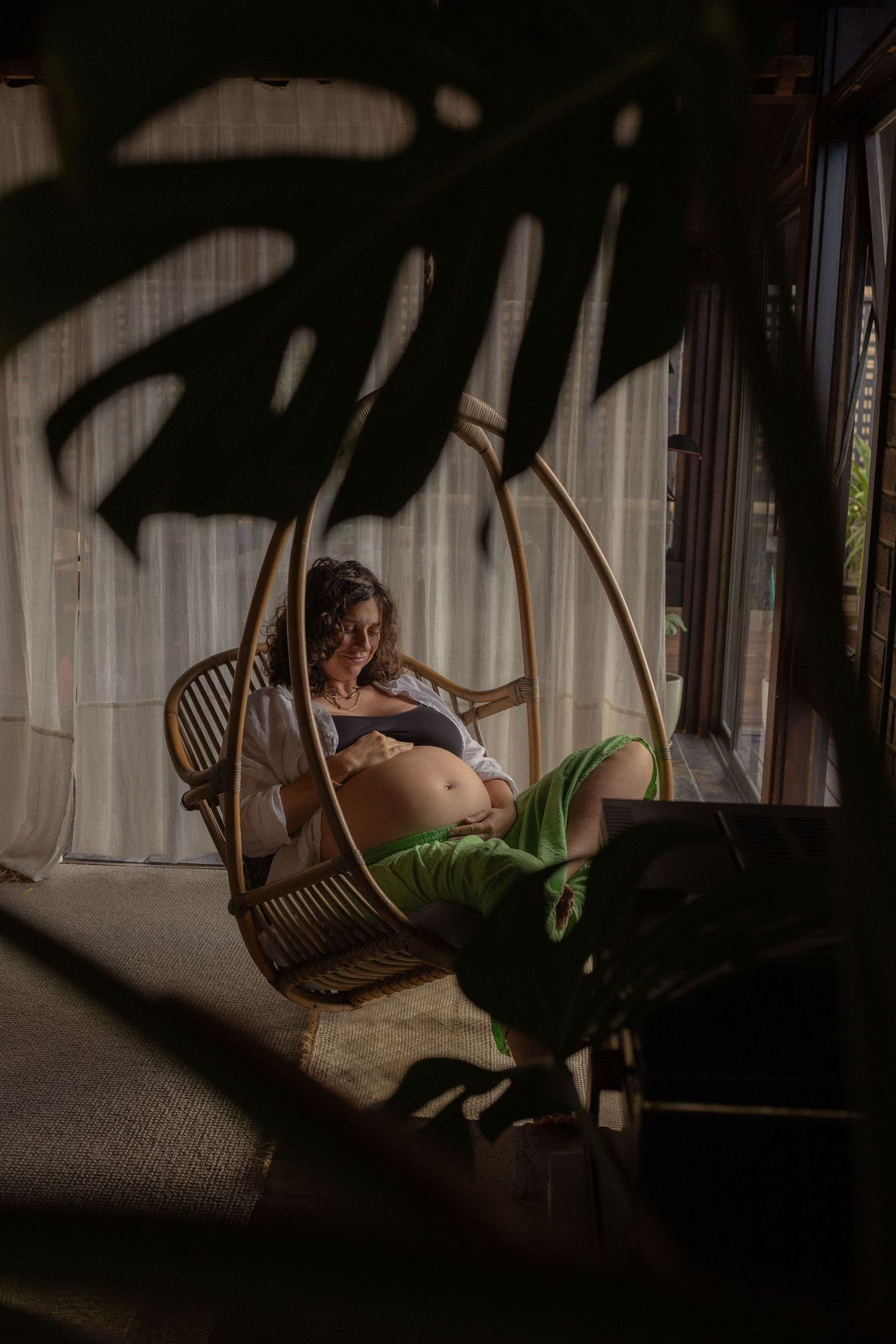 Pregnant woman sitting in a rattan hanging chair, cradling her baby bump in a sunlit room with monstera plants.