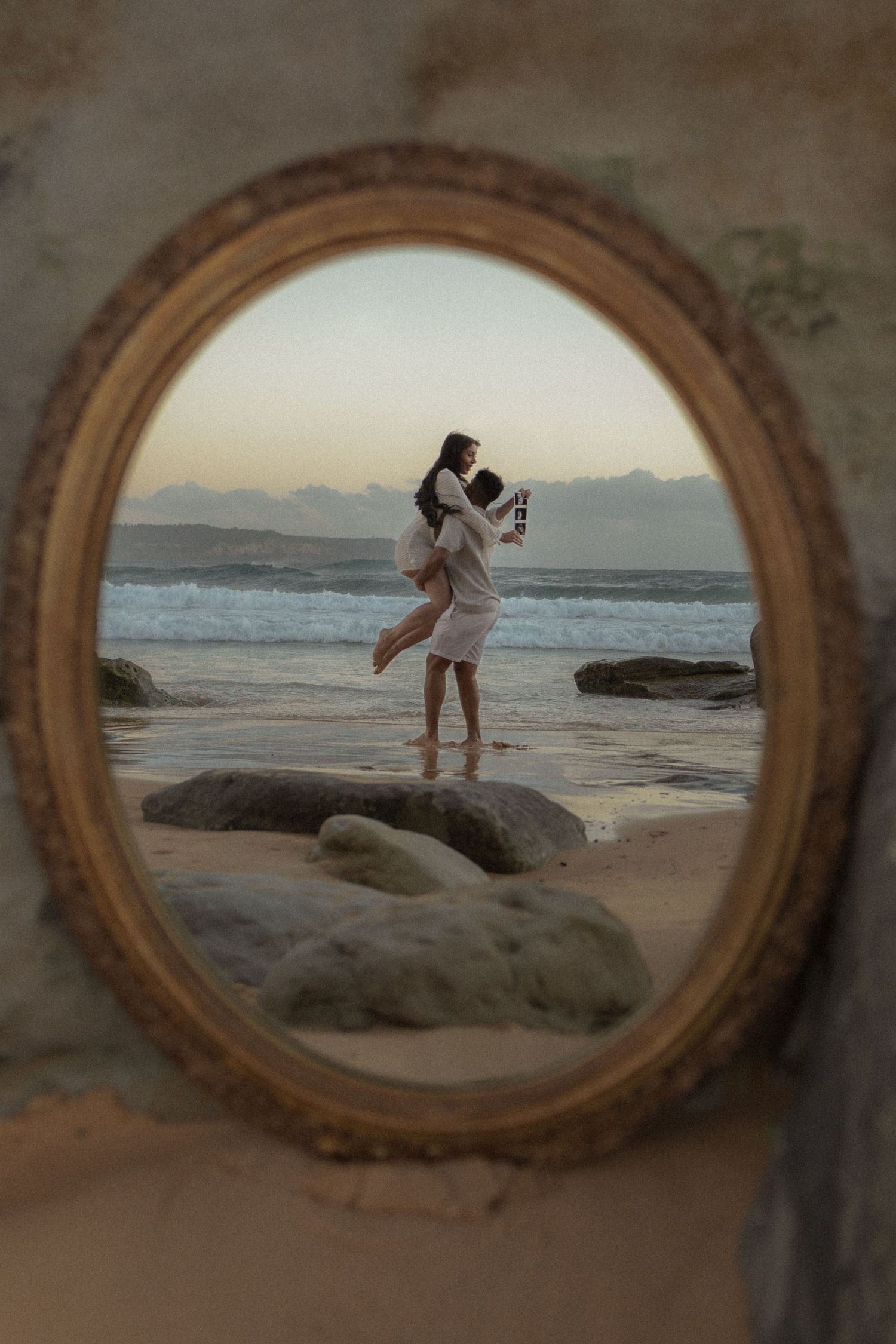Oval gold mirror on sandy beach reflecting couple embracing at ocean shore during sunset.