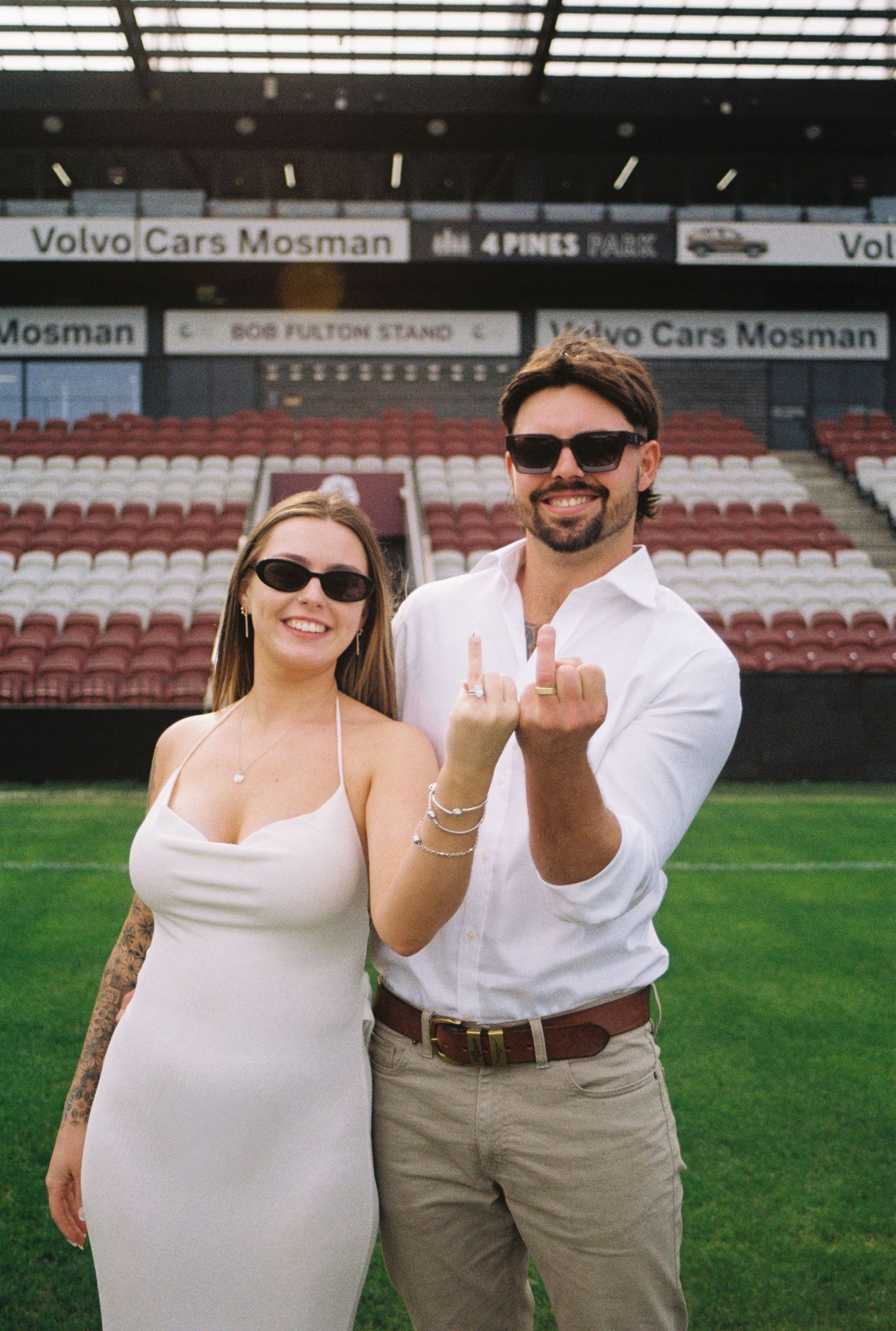 Couple smiling and showing off engagement rings on a football field stadium with red seats in the background.