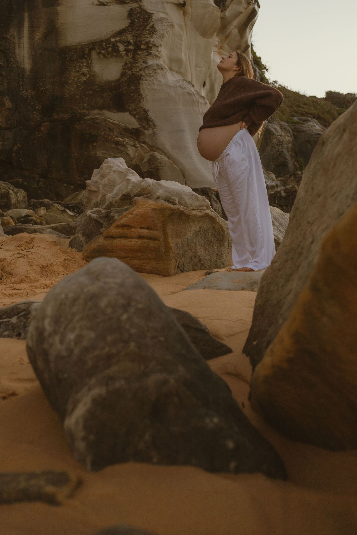 Pregnant woman in white dress and brown hat poses among sandy rocks and cliffs at golden hour.