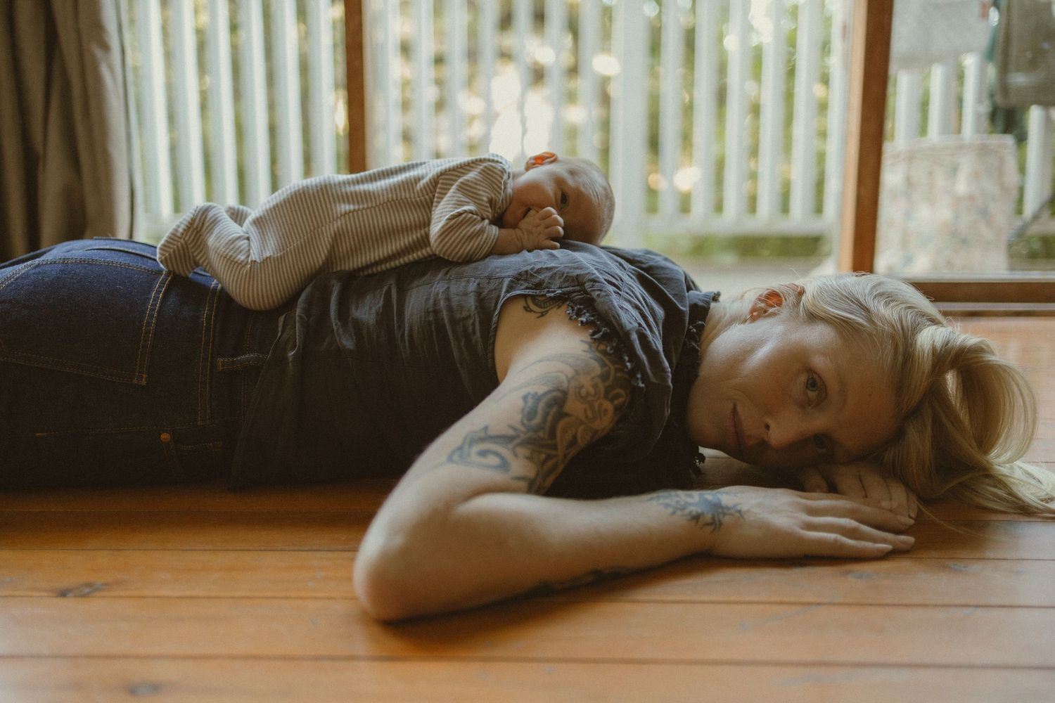 Tattooed blonde woman lying on hardwood floor in denim outfit, resting head on arms near large windows.
