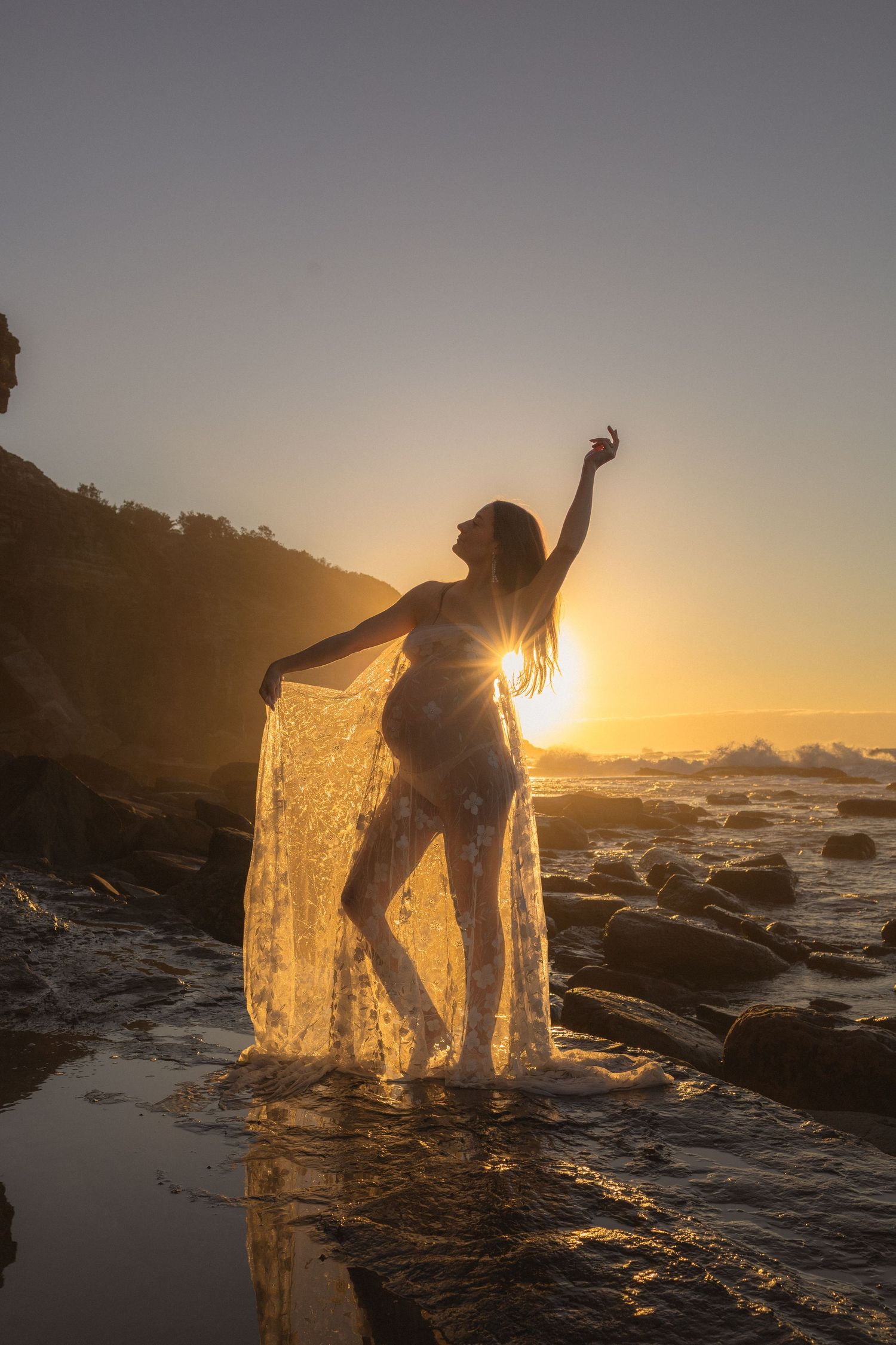 Pregnant woman in lace maternity gown dancing joyfully on rocky beach at golden sunset.