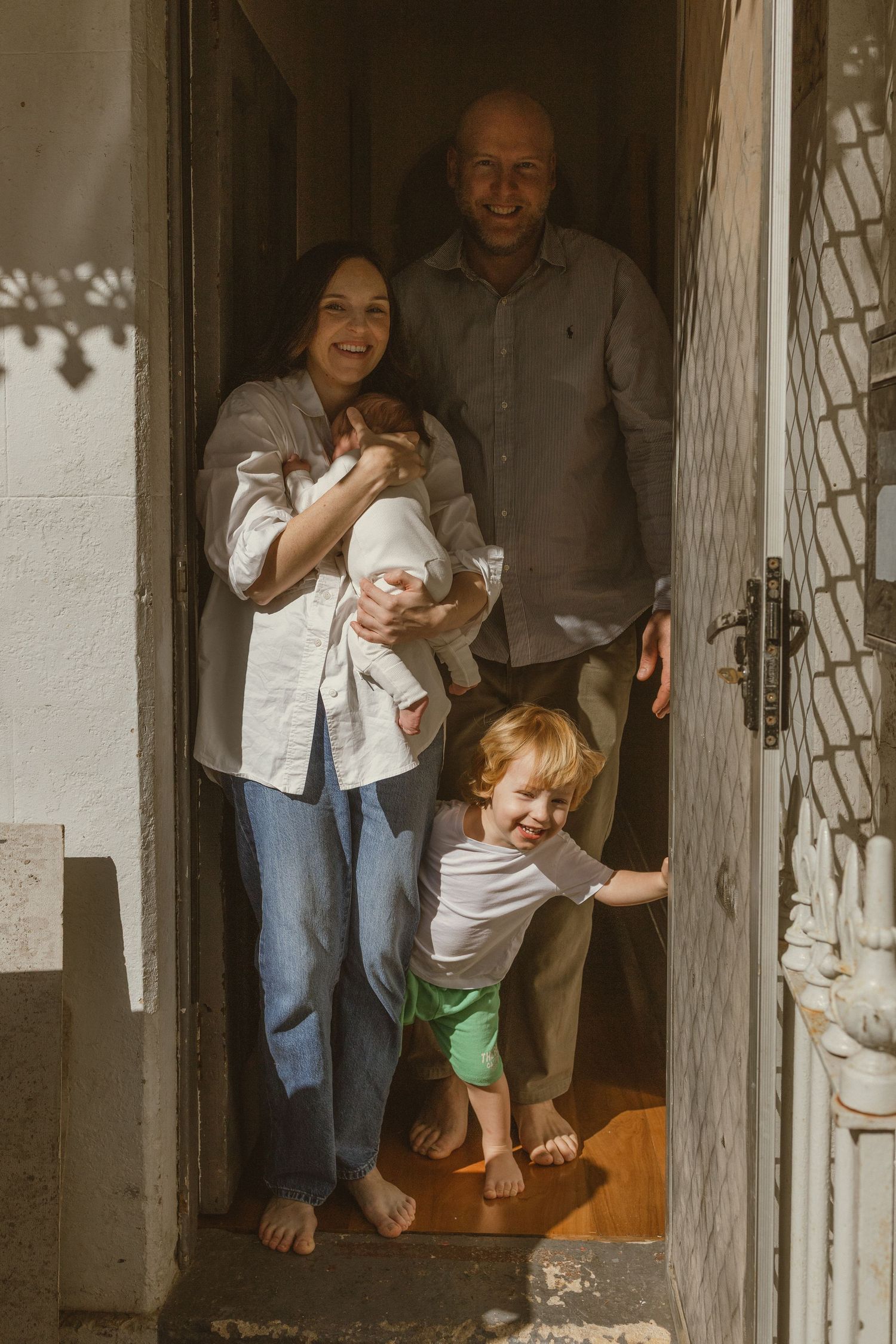 Happy family of four with newborn baby and toddler posing in sunlit doorway of their home.