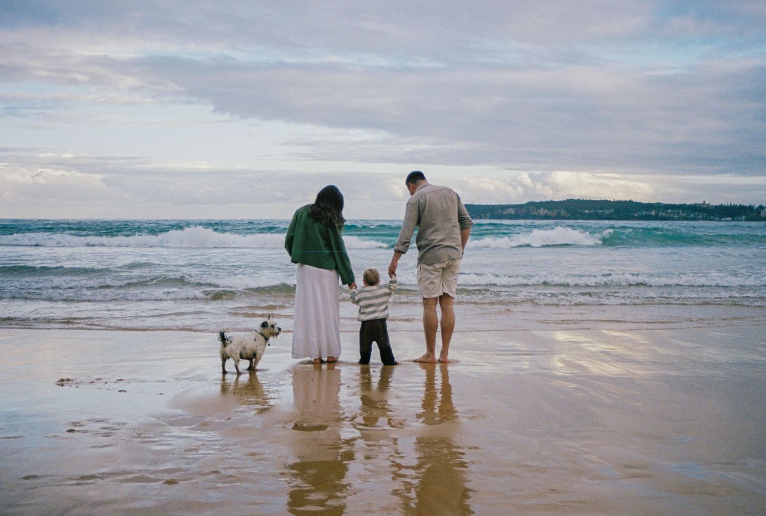 Family with toddler and small dog standing on sandy beach watching ocean waves, rear view on cloudy day.
