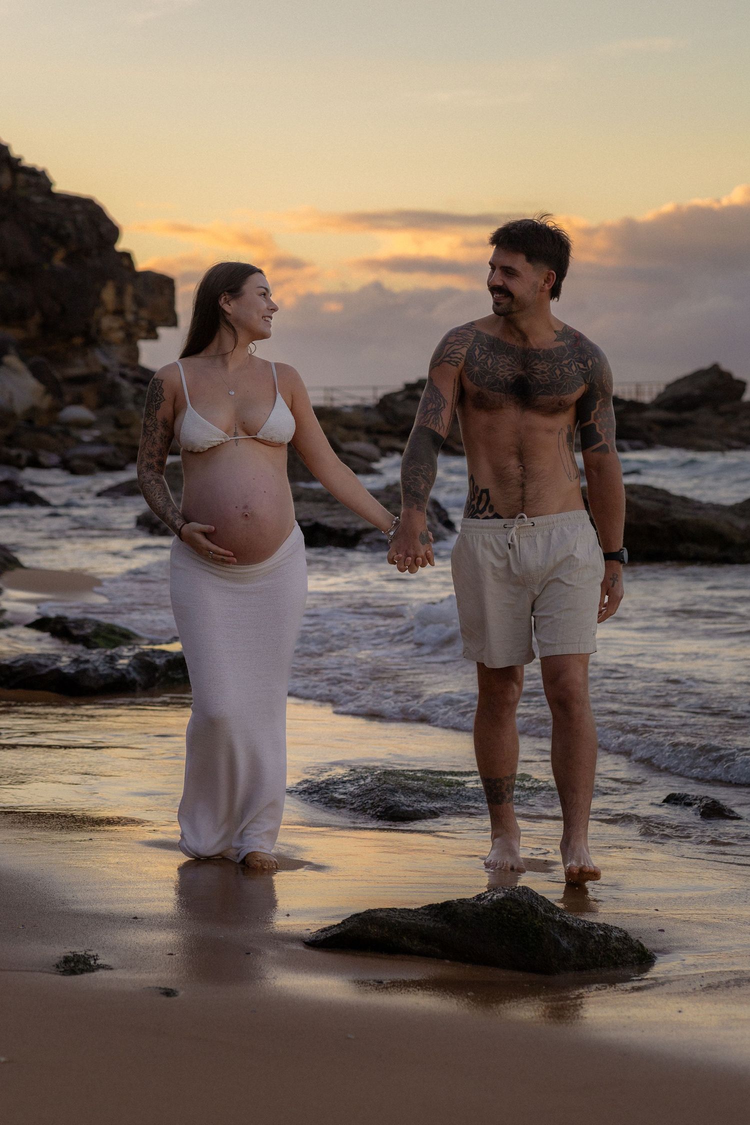 Tattooed couple holding hands on a beach at sunset, pregnant woman in white dress cradling baby bump.