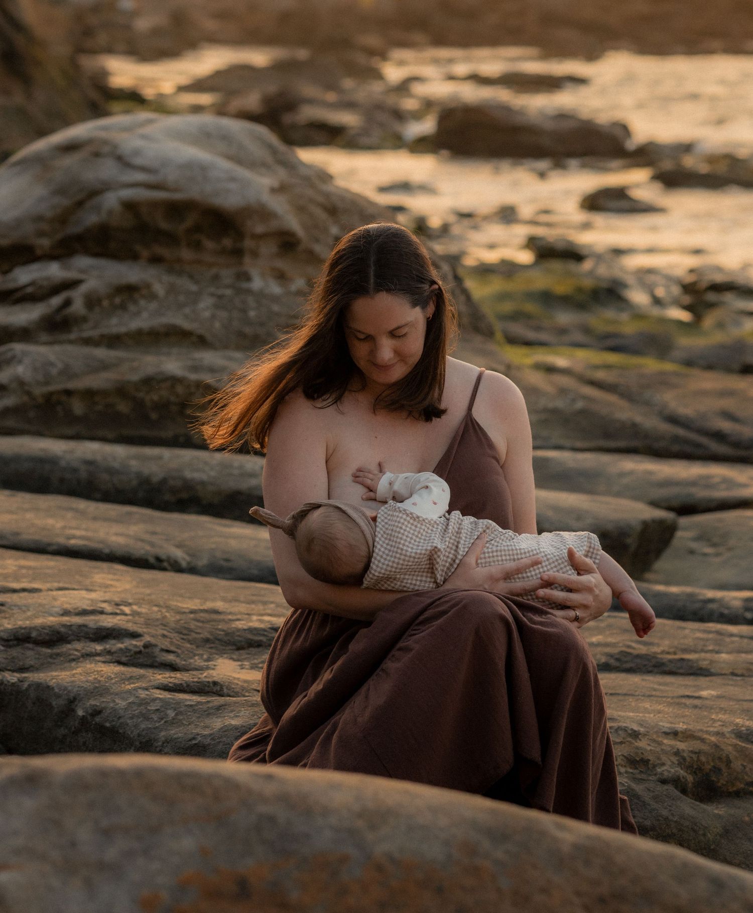 A mother nursing her infant while sitting on rocky coastal terrain at sunset in warm golden light.
