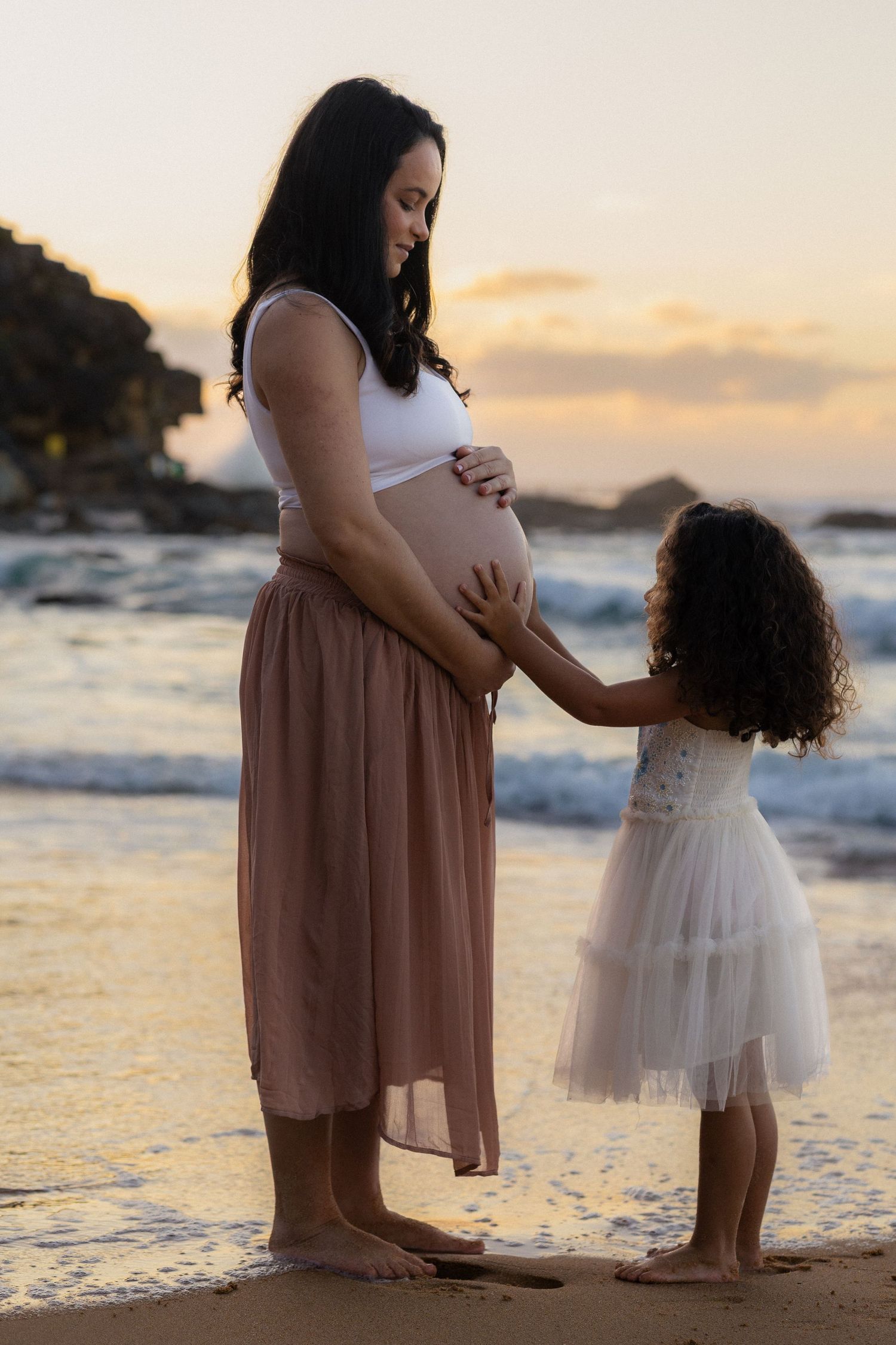 A mother and daughter share a tender moment at sunset on a beach with gentle waves in the background.