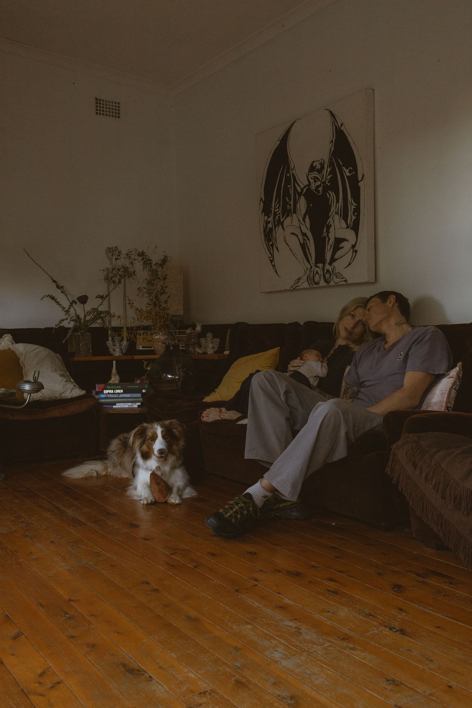 Couple relaxing on dark sofa with Cavalier King Charles Spaniel on hardwood floor, gothic angel artwork on wall.