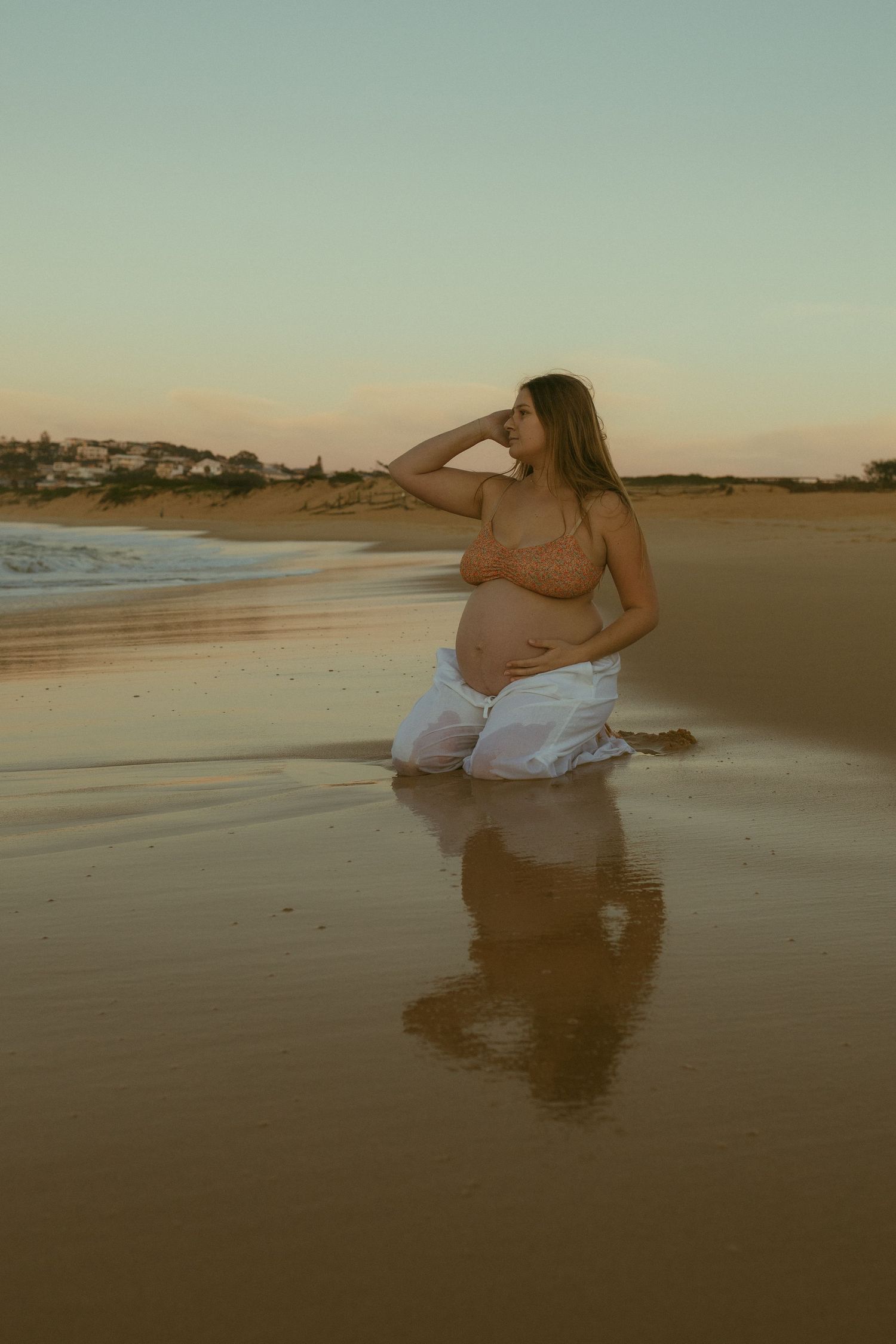 Pregnant woman kneeling on sandy beach at sunset, wearing white wrap, hand shading eyes, with ocean waves behind her.