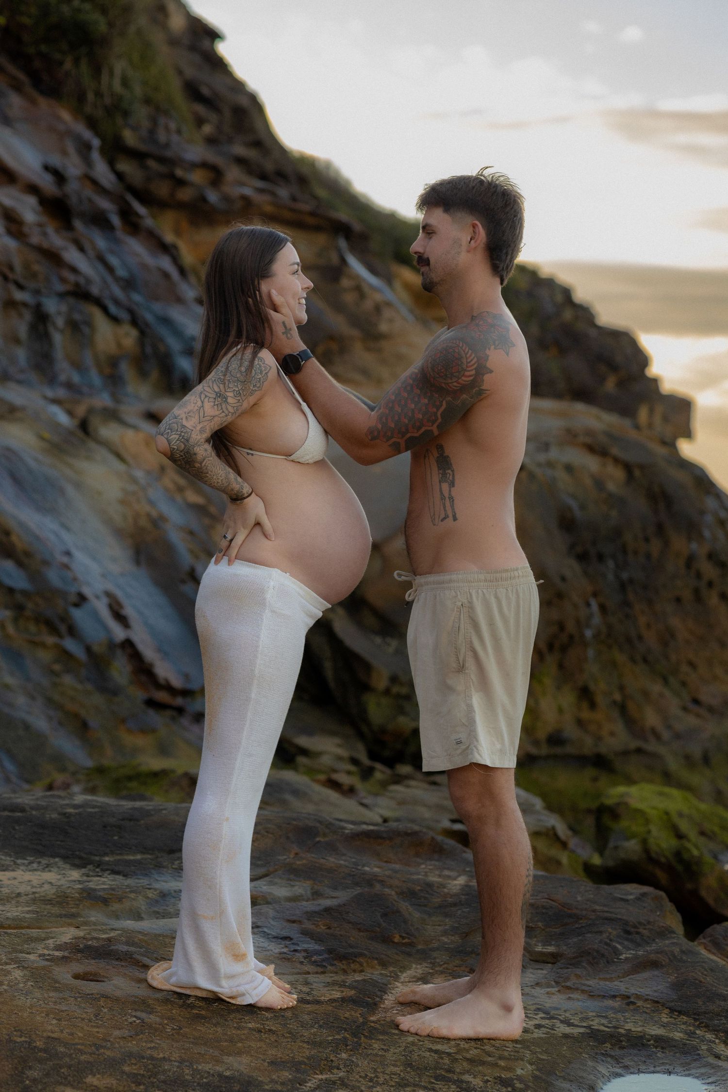 Tattooed pregnant couple standing on coastal rocks at sunset, man tenderly holding woman's face in a romantic maternity photo.