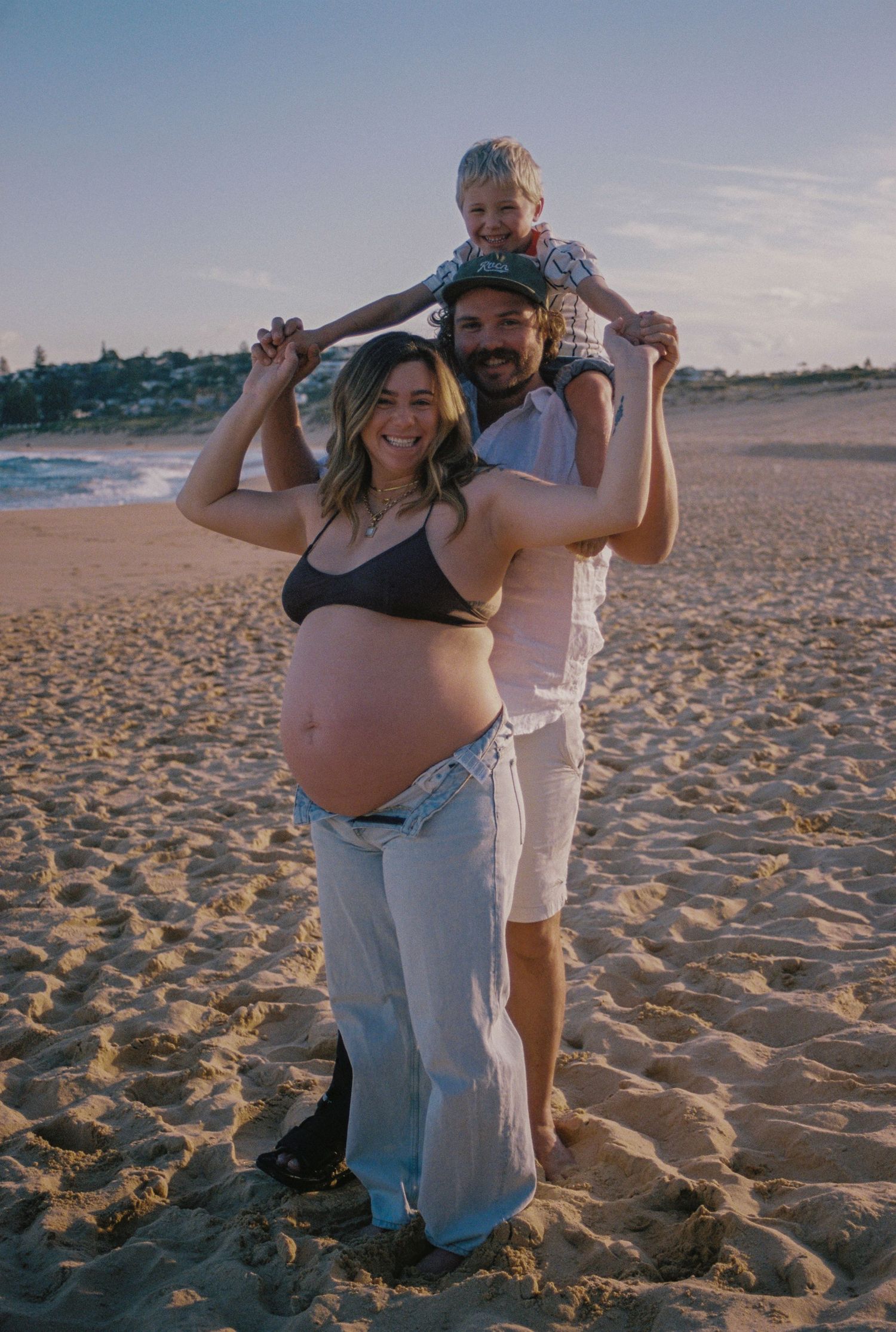 Pregnant woman poses with partner and child on sandy beach at sunset during maternity photo shoot.