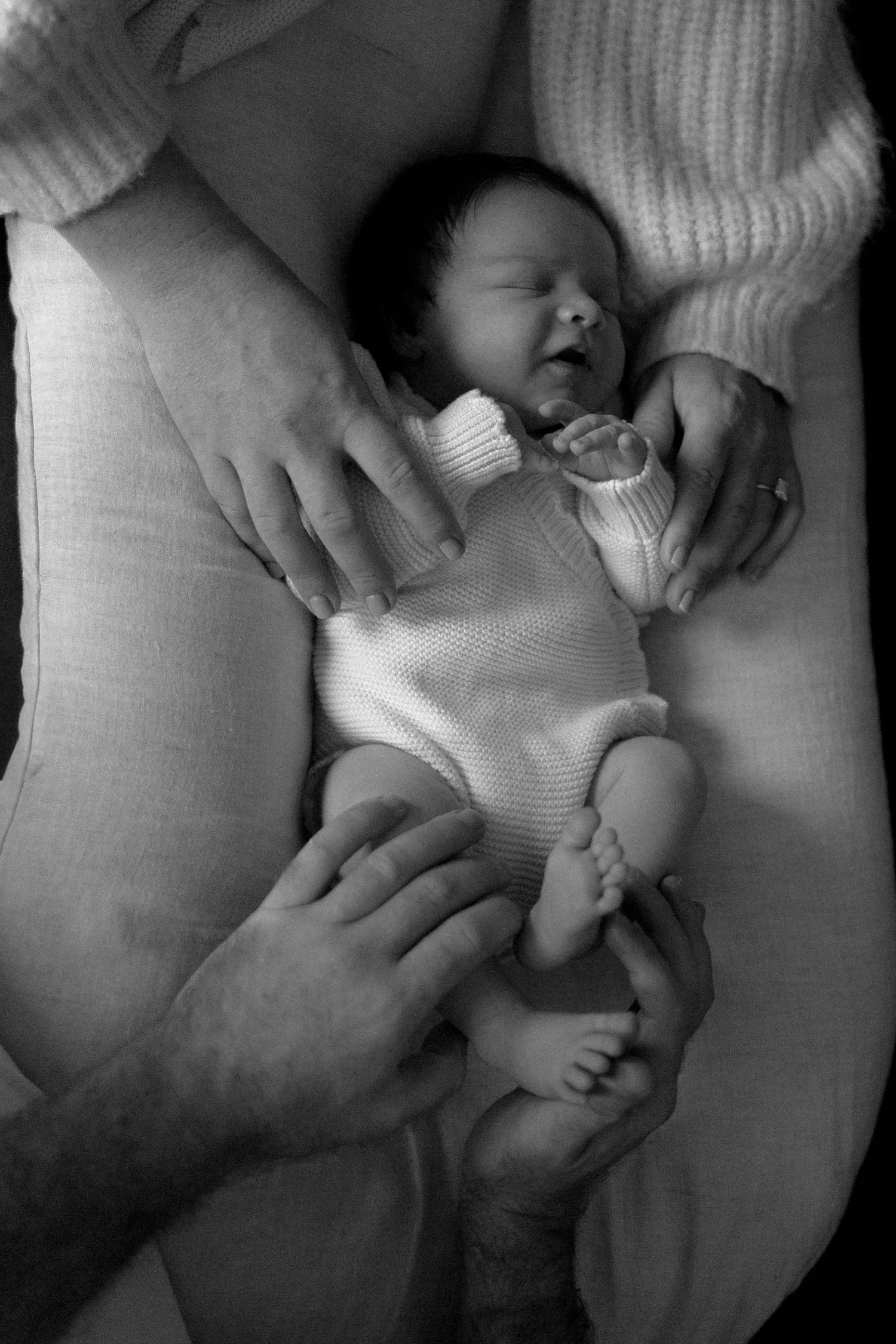 Black and white photo of a sleeping newborn baby cradled in parents hands, wearing a white knit outfit.
